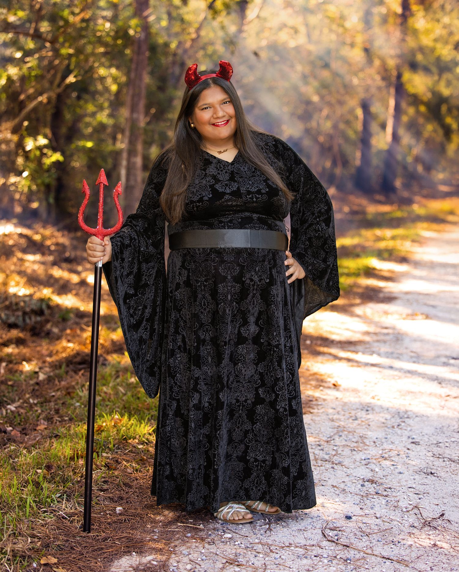Outdoor photoshoot featuring a black velvet dress and red devil's pitchfork prop on a sunlit forest path.