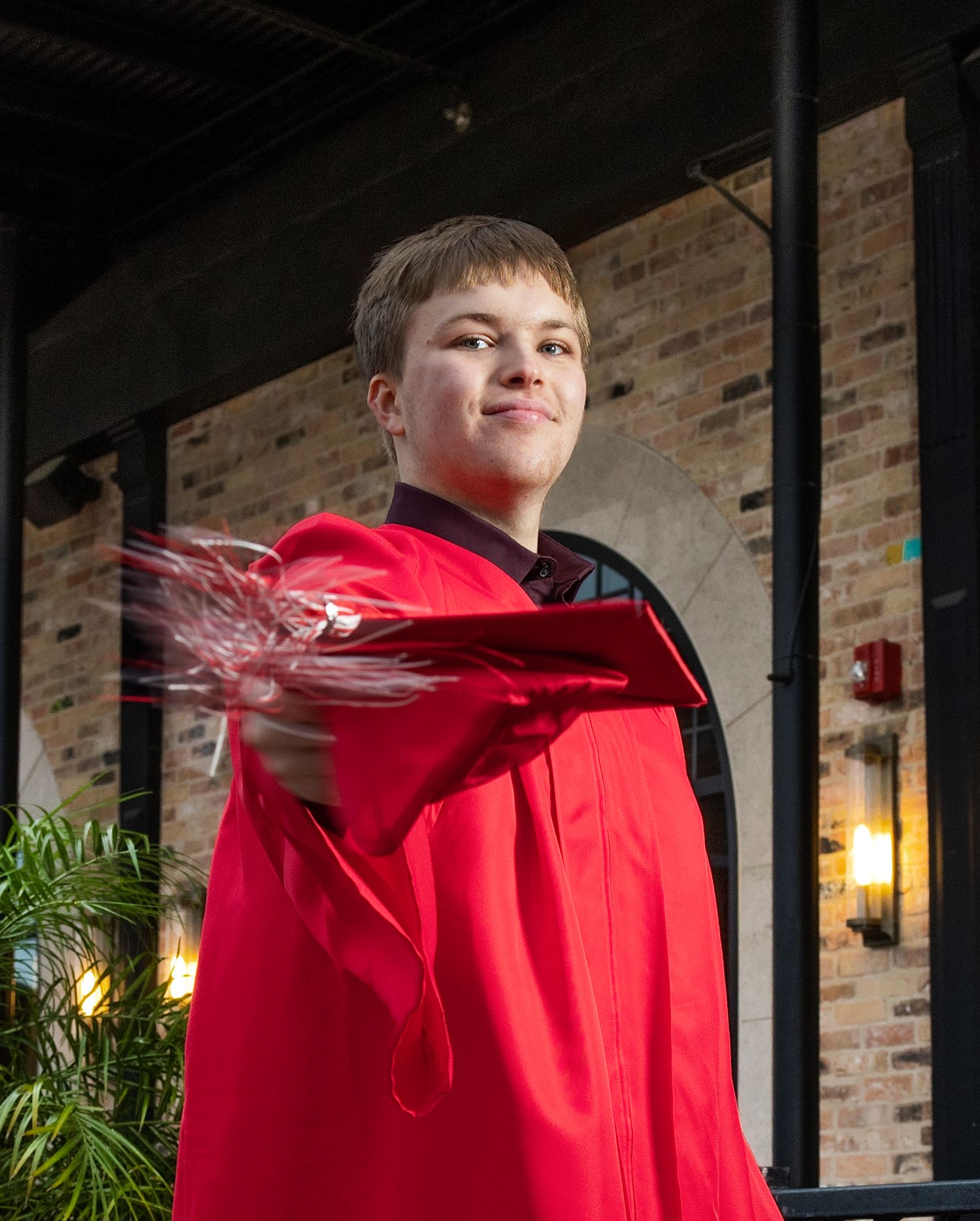 A person in a bright red hoodie stands against a brick wall with black vertical bars in an indoor setting.