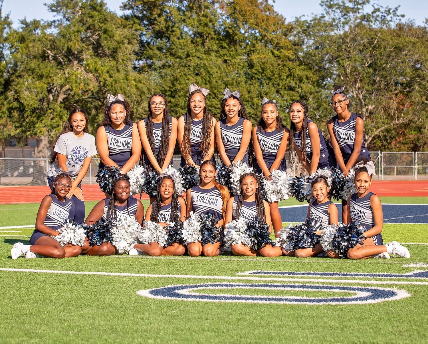 Cheerleading team sits in formation on a sports field with trees in the background during a sunny day.
