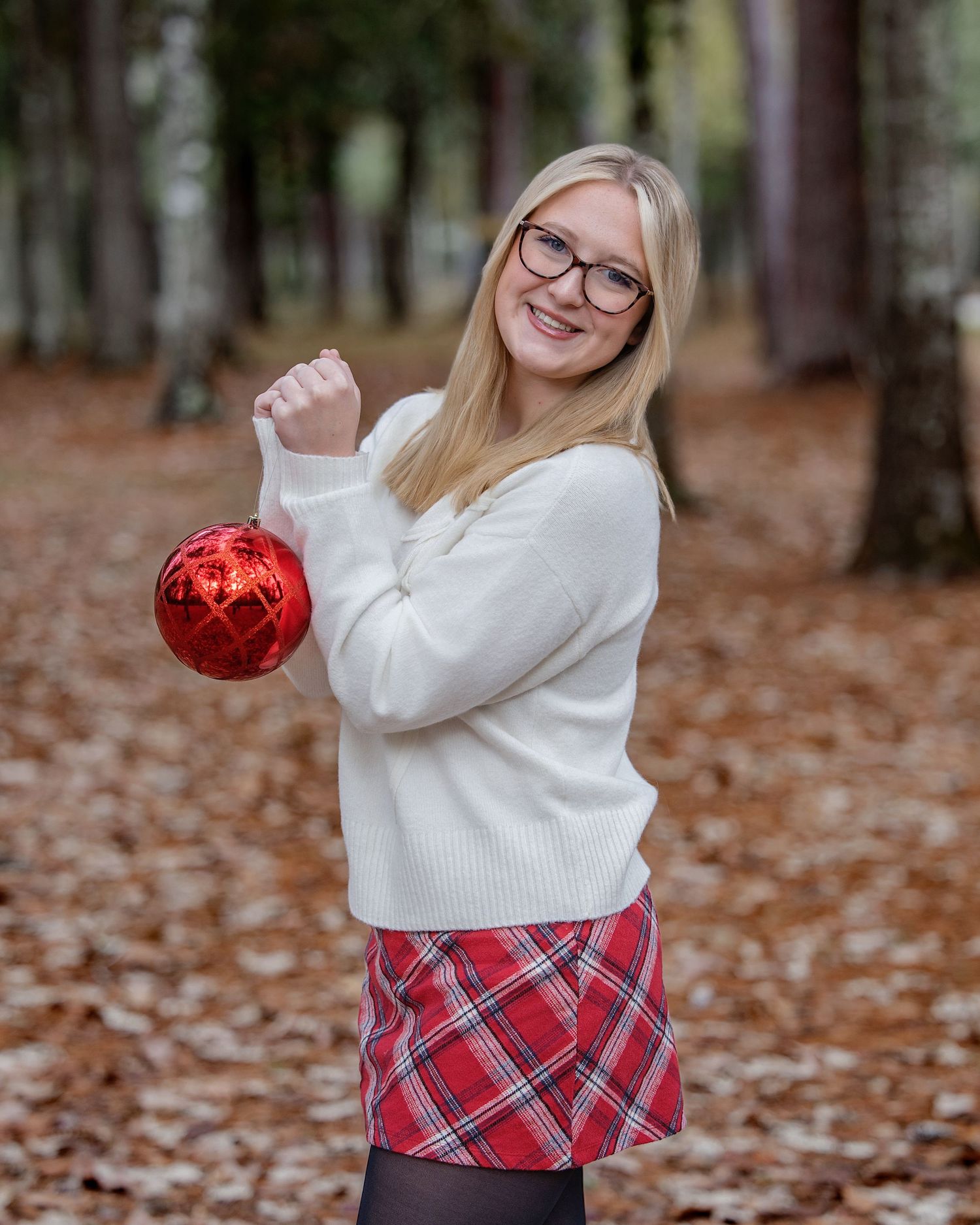 Someone wearing a white sweater and plaid skirt poses playfully with a red ornament among fallen autumn leaves.