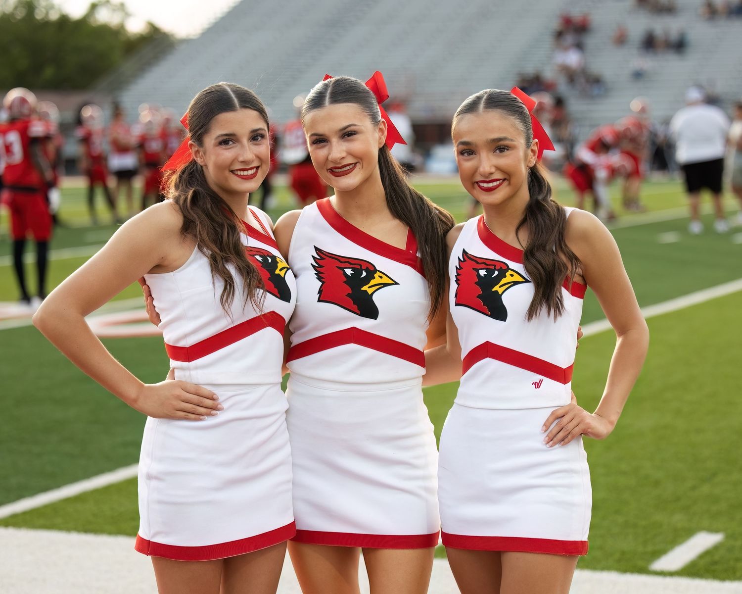 Cheerleaders in white and red uniforms pose together on a football field during a game day performance.