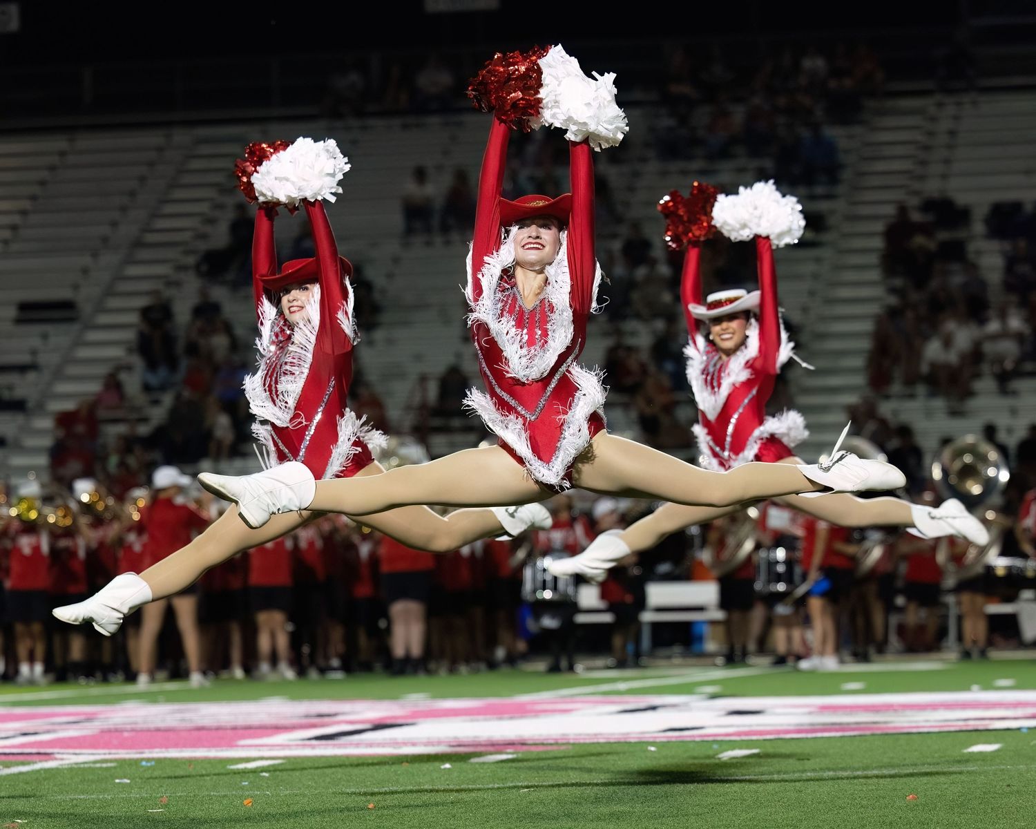 Cheerleaders perform synchronized jumps in red and white uniforms during a nighttime football game.