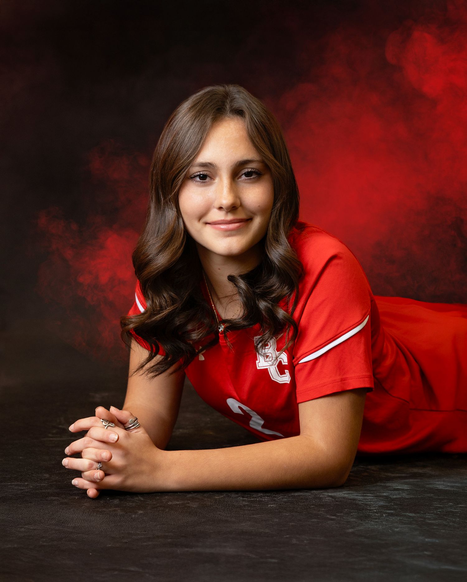 Athletic portrait against dramatic red and black backdrop featuring a person in a red sports jersey lying on the ground.