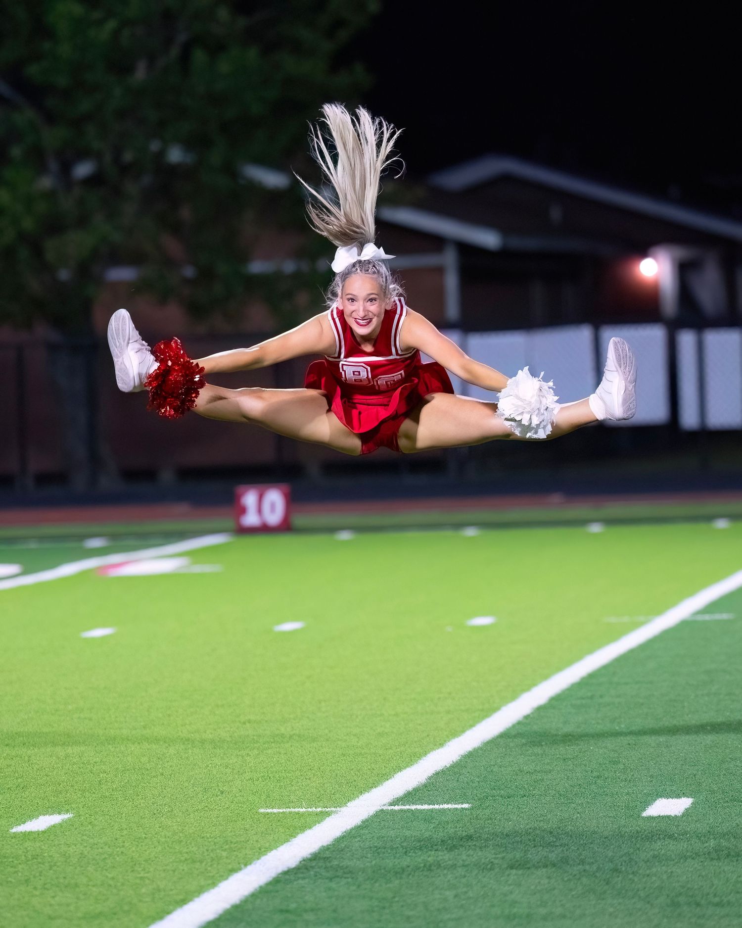 Cheerleader performs a mid-air split jump in red uniform on football field at night.