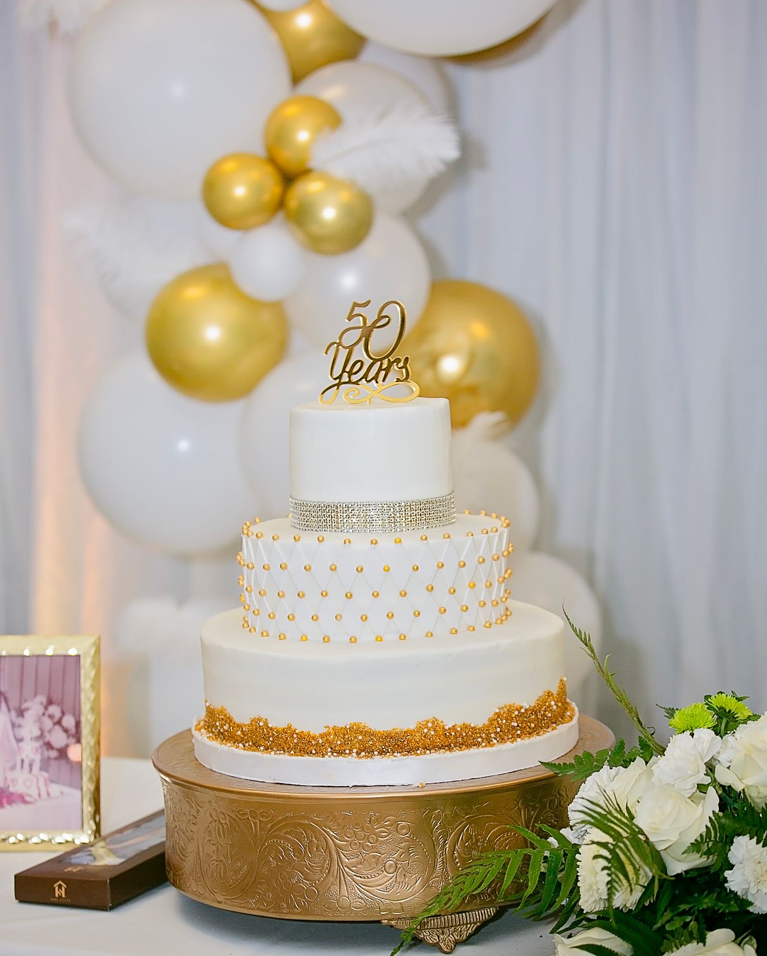 Elegant white and gold 50th anniversary celebration cake with balloon decorations and floral arrangements.