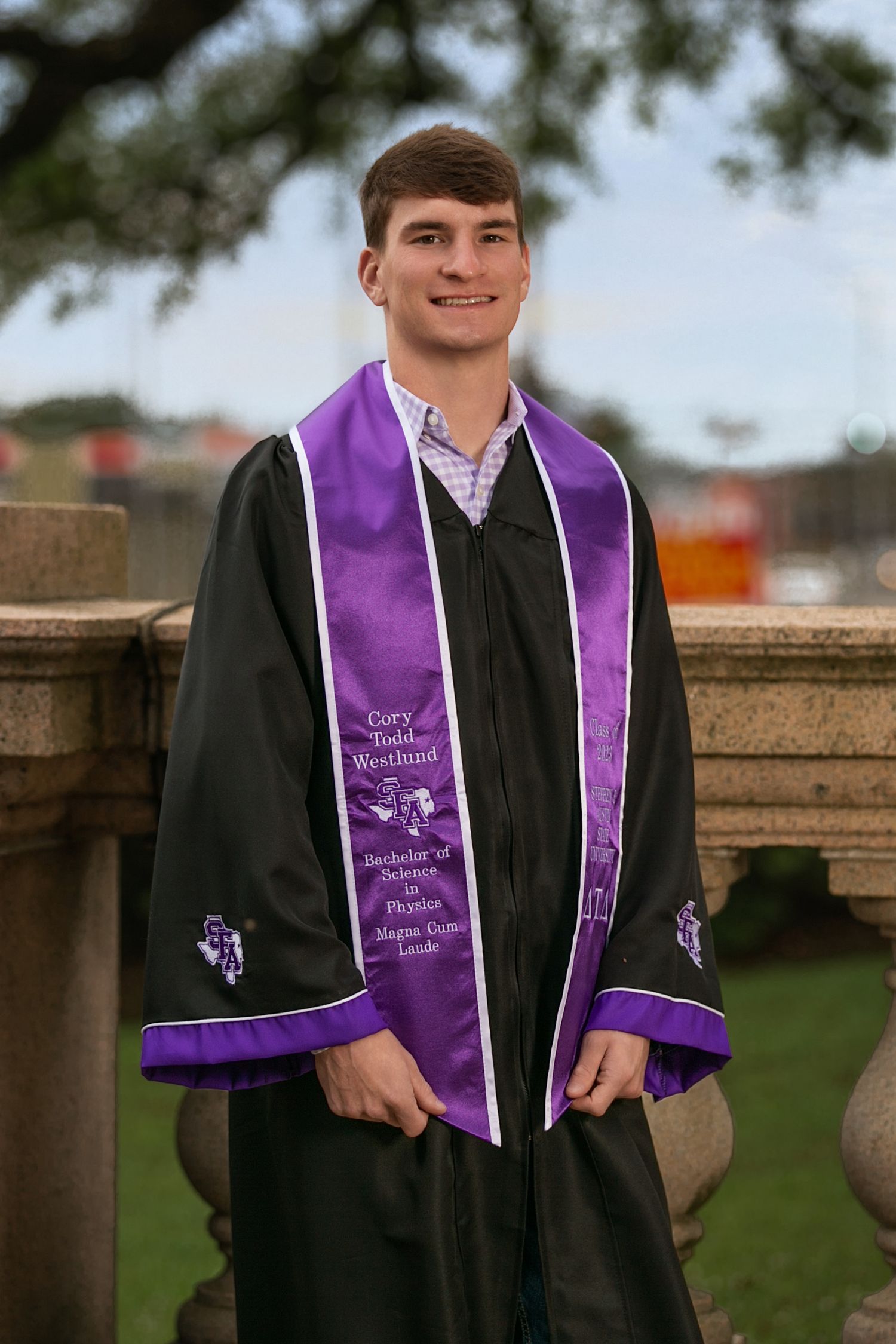 College Graduate wearing black gown with purple stole and sash stands outdoors near wooden fence during graduation photo session.