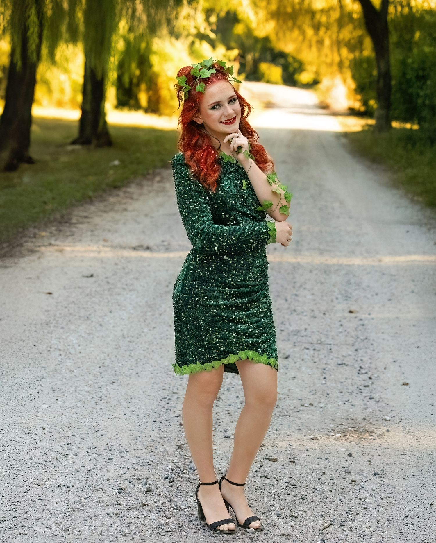 A person in a sparkly green dress poses playfully on a sunlit gravel path surrounded by trees.