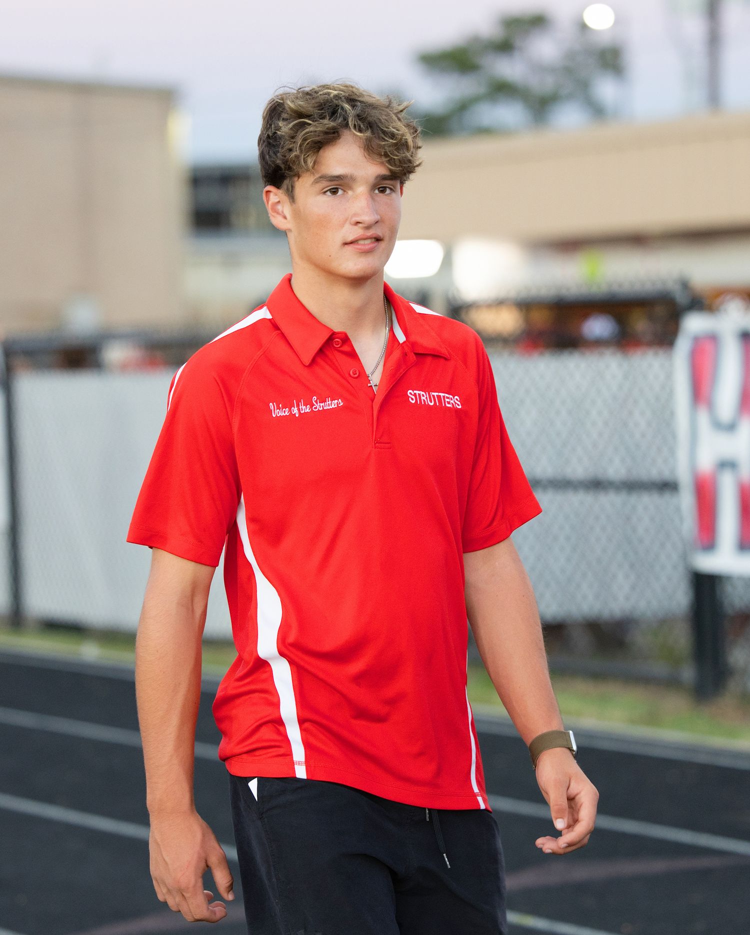 Person in red polo shirt and black pants walking along track at sports facility.