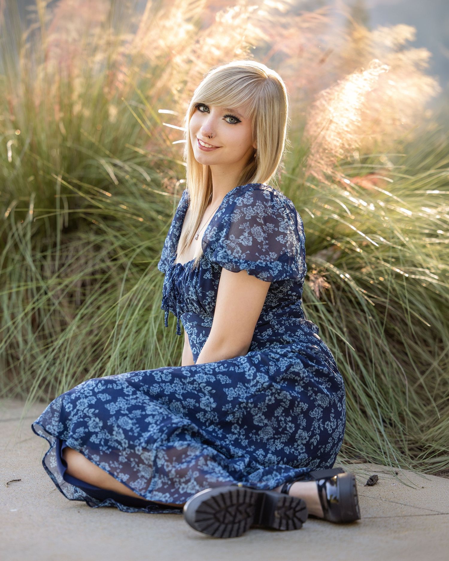 Young woman with blonde hair sitting outdoors in a blue floral dress, smiling against tall ornamental grasses.