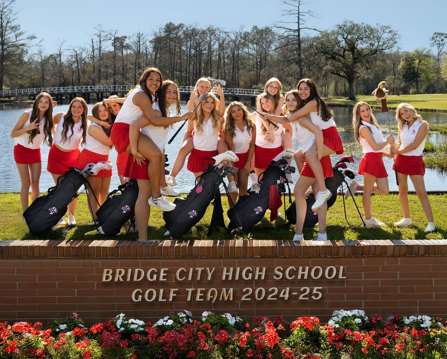 High school golf team poses playfully with their golf clubs on a brick wall near a lake in red and white uniforms.