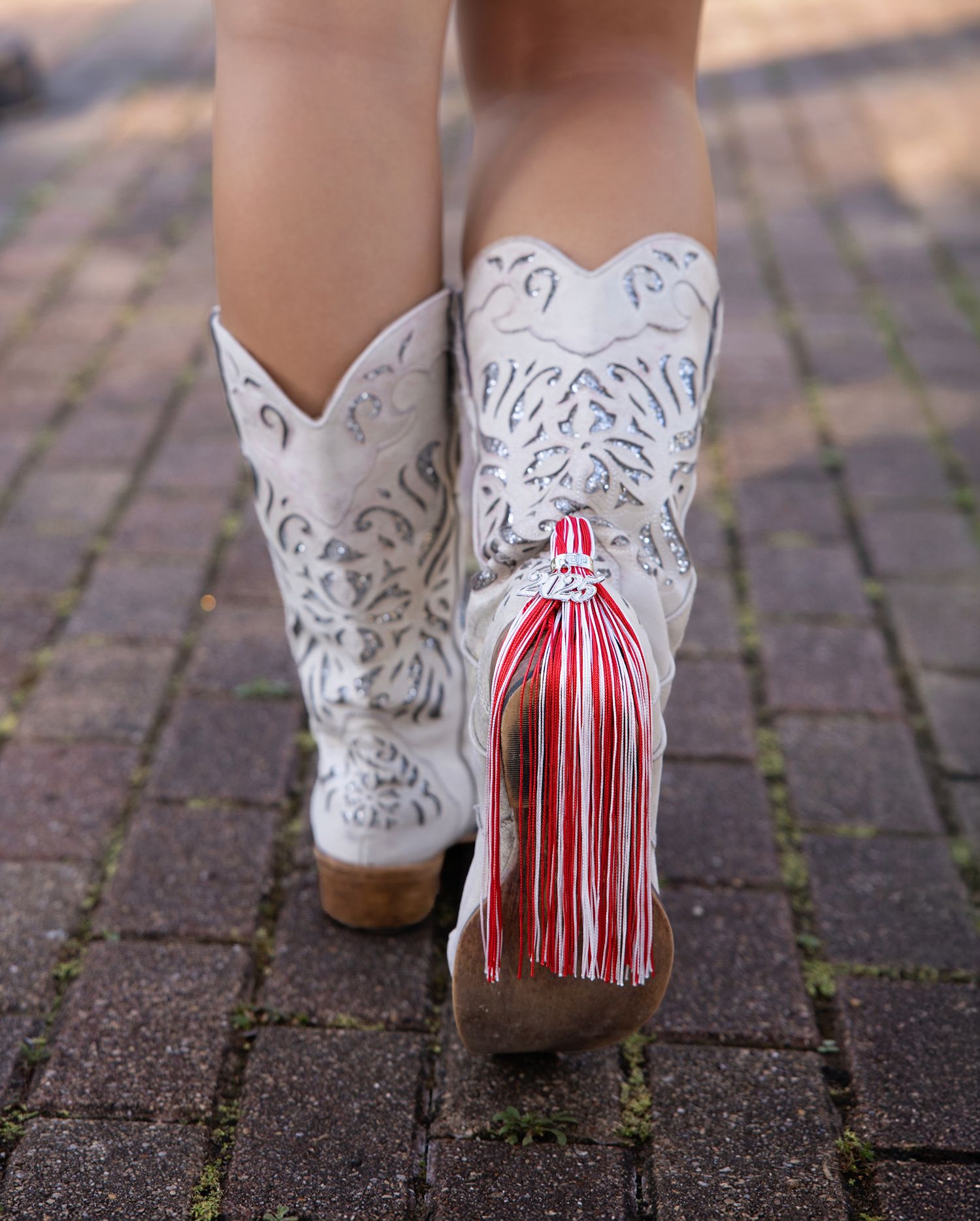 White cowboy boots with decorative cutouts and red and white striped tassels on brick pavement.