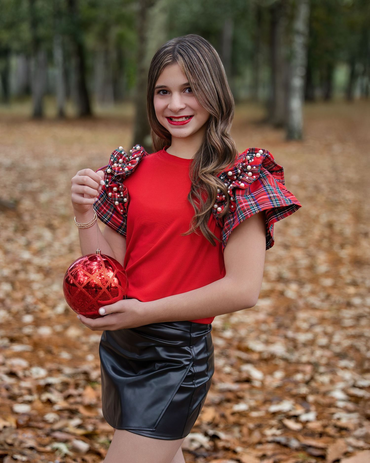 Fashion model wearing red top and black leather skirt poses playfully in autumn forest setting with fallen leaves.