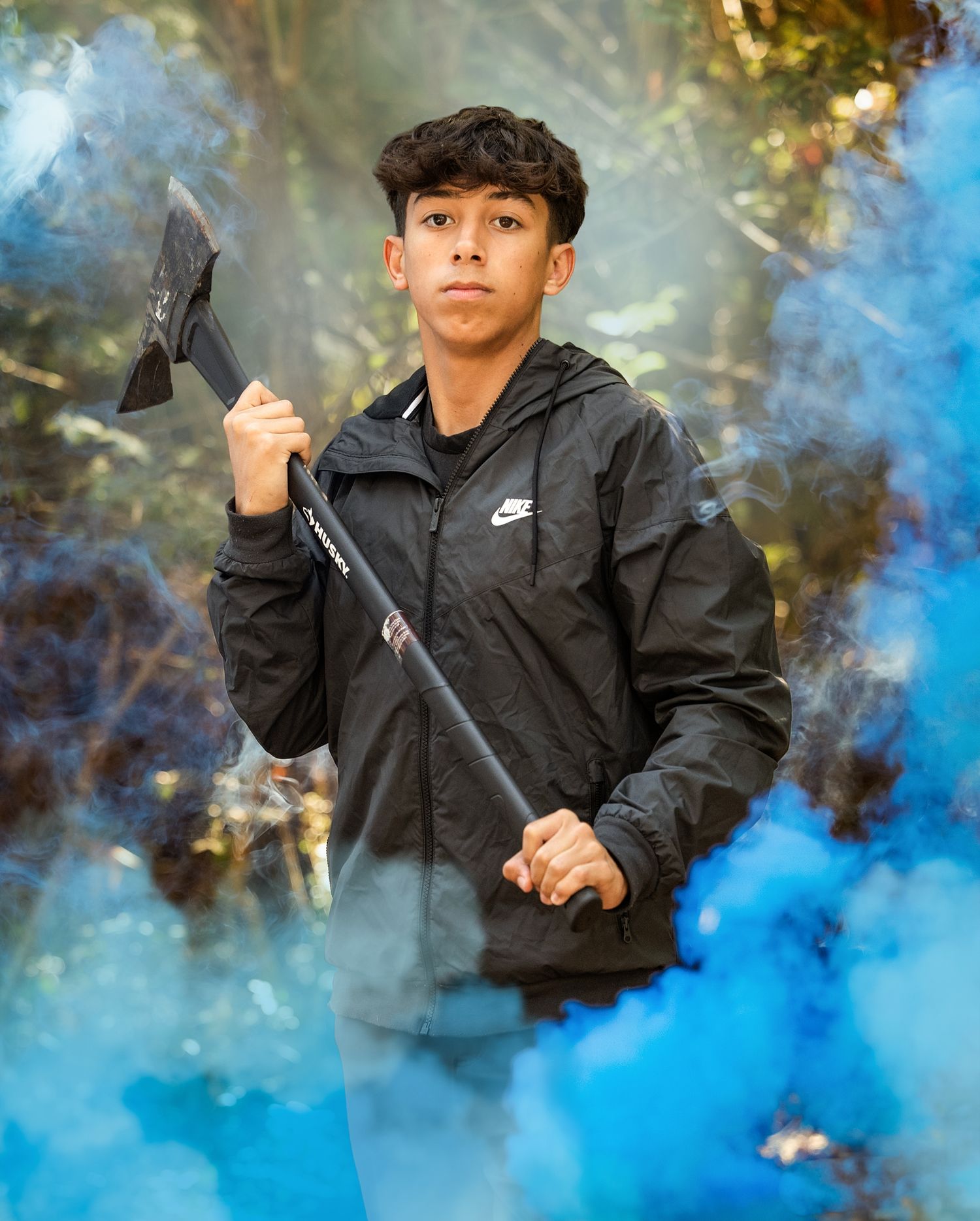 A person in a Nike jacket holds an axe prop while surrounded by dramatic blue smoke effects outdoors.