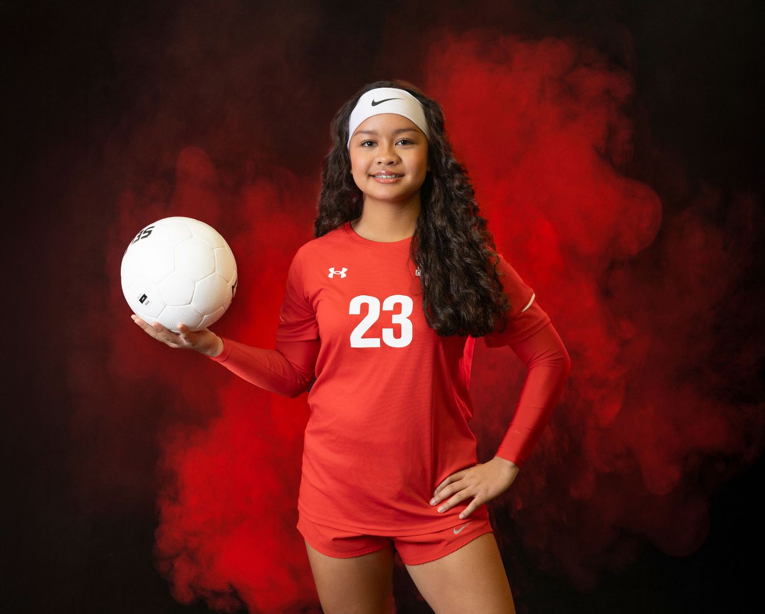 Volleyball player in red uniform poses with ball against dramatic red smoke background for sports portrait session.