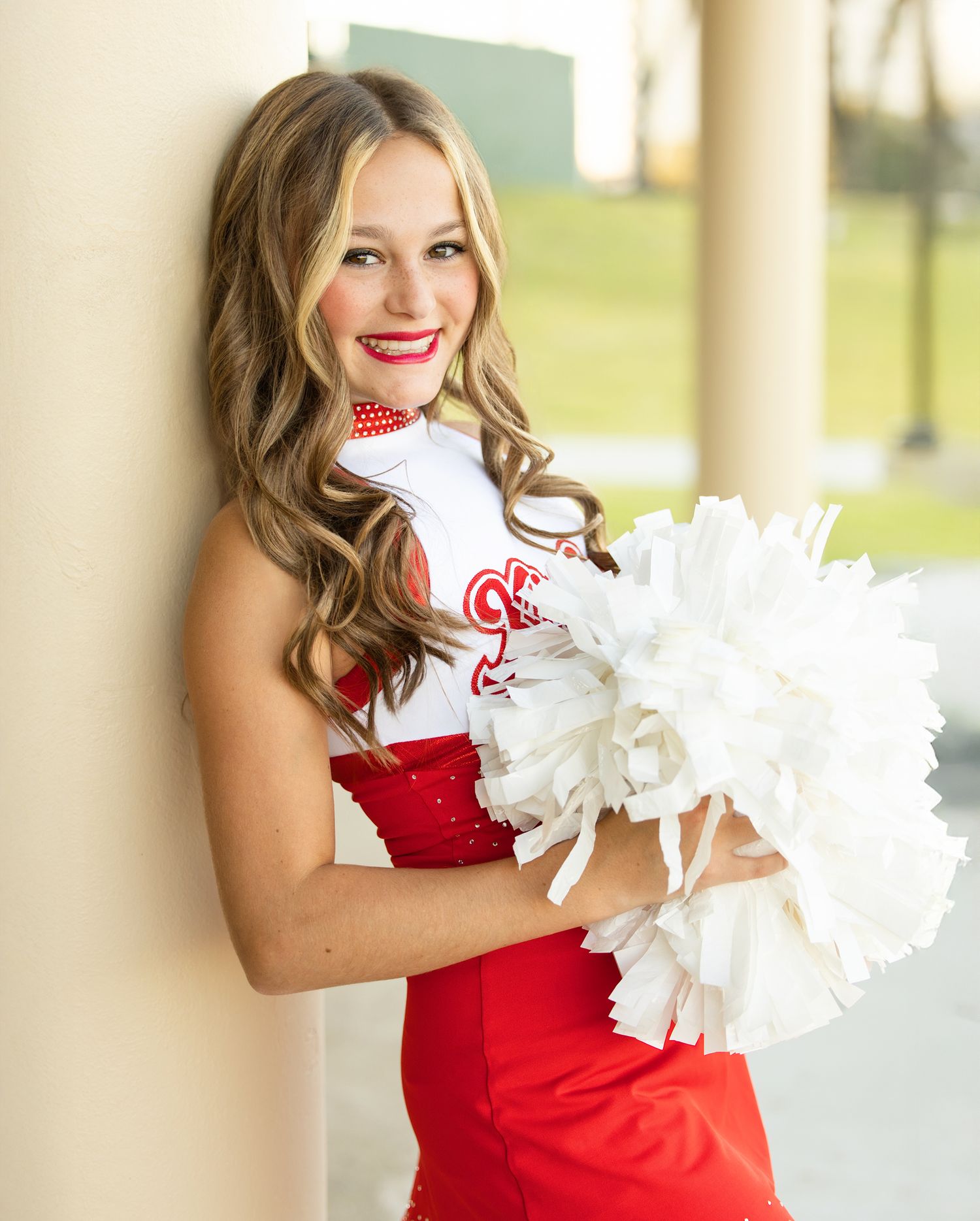 Cheerleader in red uniform holds white pom poms while posing against wall.