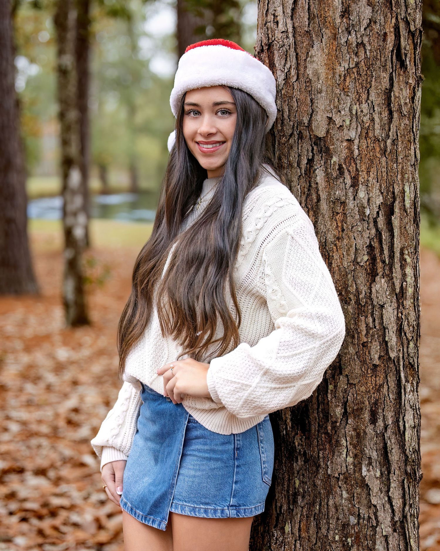 Laney a beautiful Vidor HS Senior in white sweater and denim skirt poses playfully by tree trunk in autumn setting with fallen leaves.