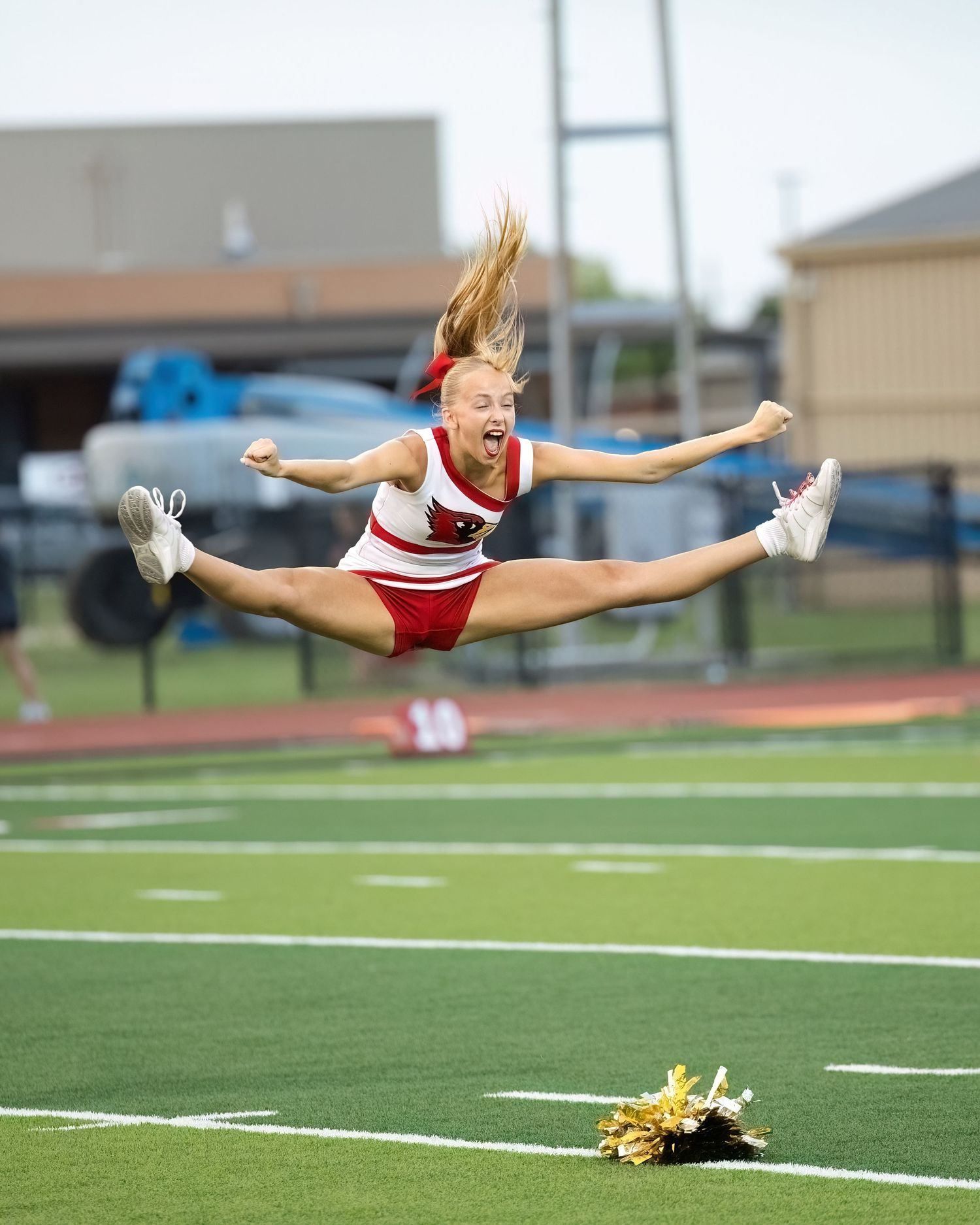 Cheerleader performs energetic toe-touch jump on football field with pom poms visible on turf below.