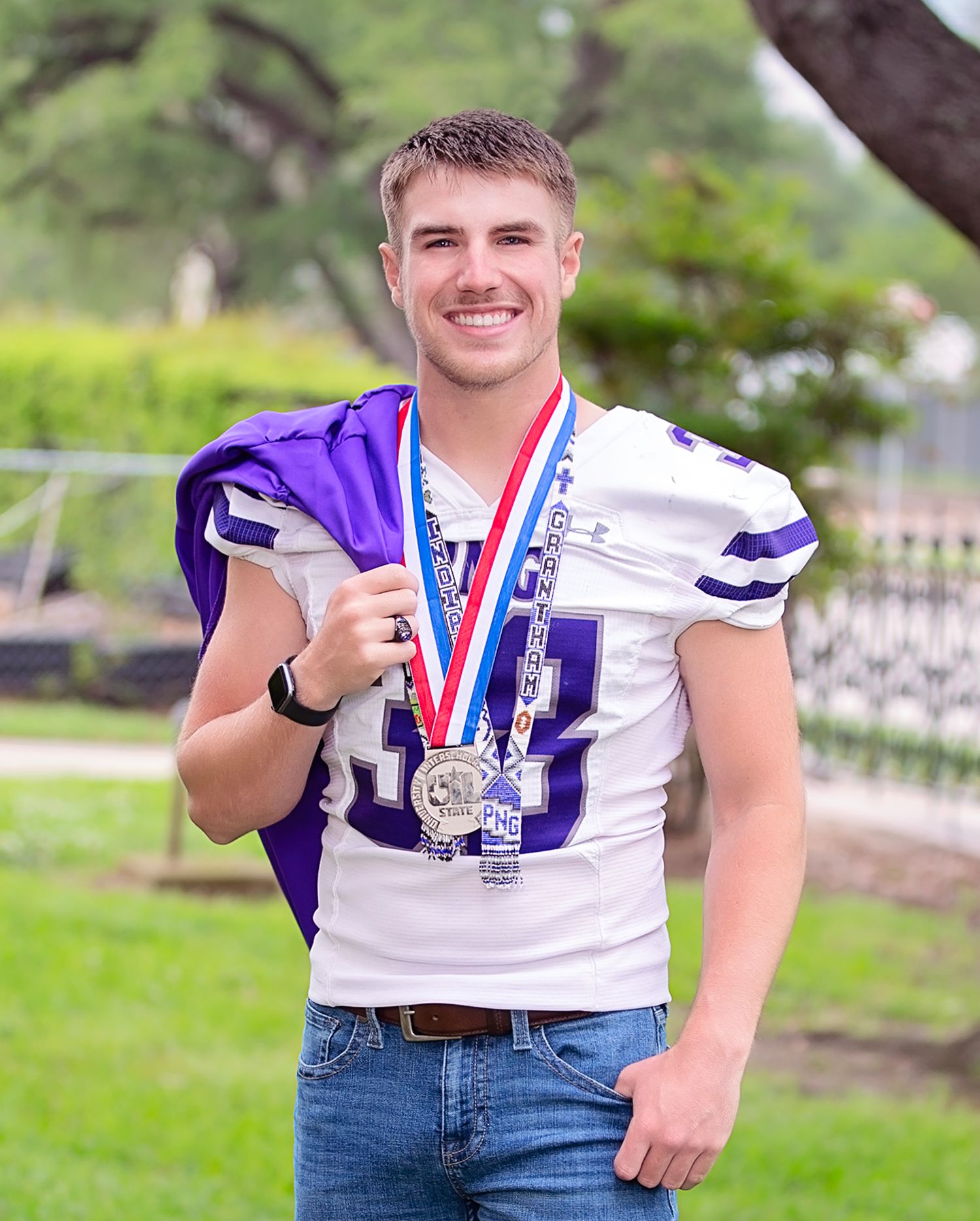 Young athlete in white football jersey proudly displays medals and purple team flag outdoors.