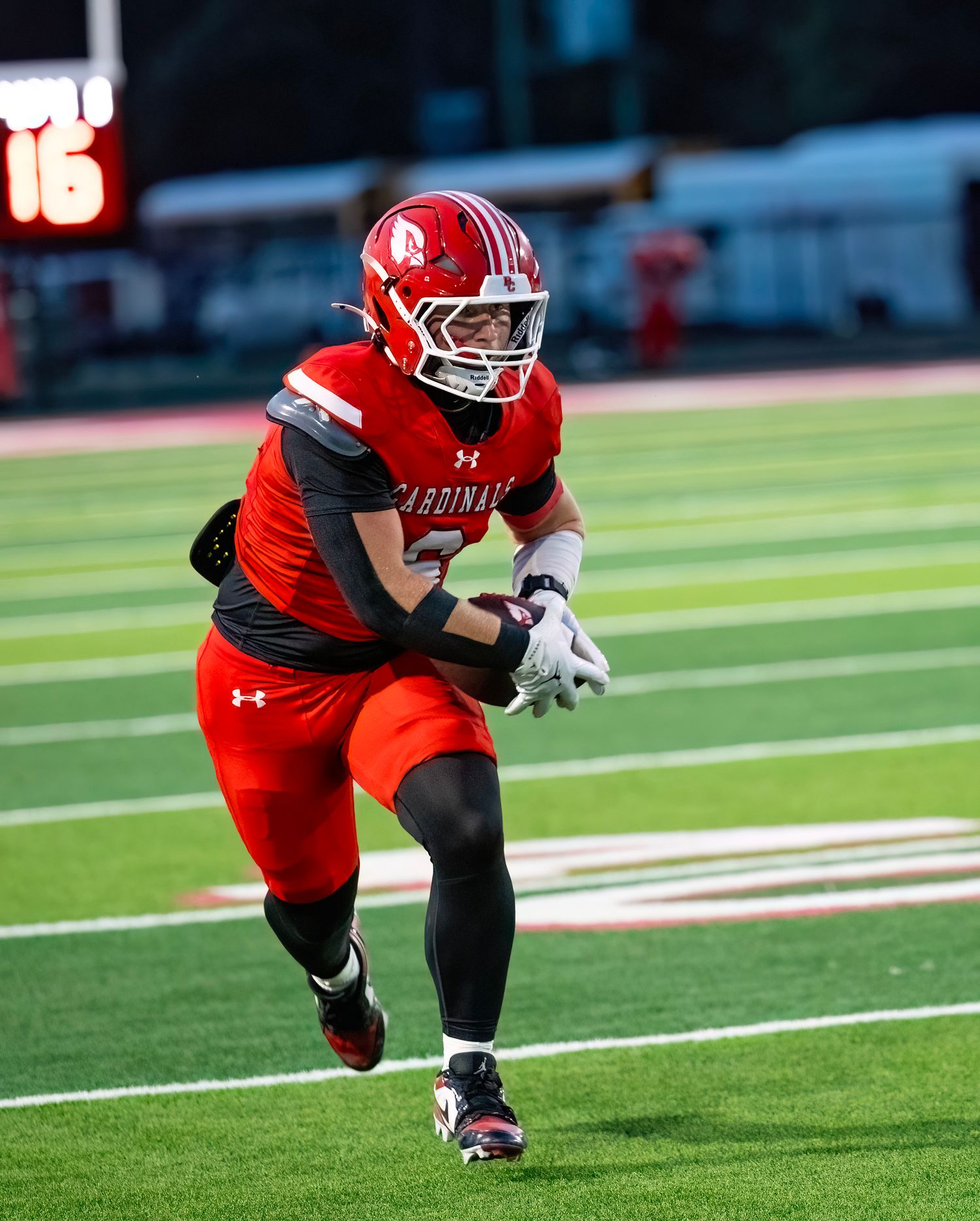 Football player in red uniform crouches low in athletic stance on field during game.