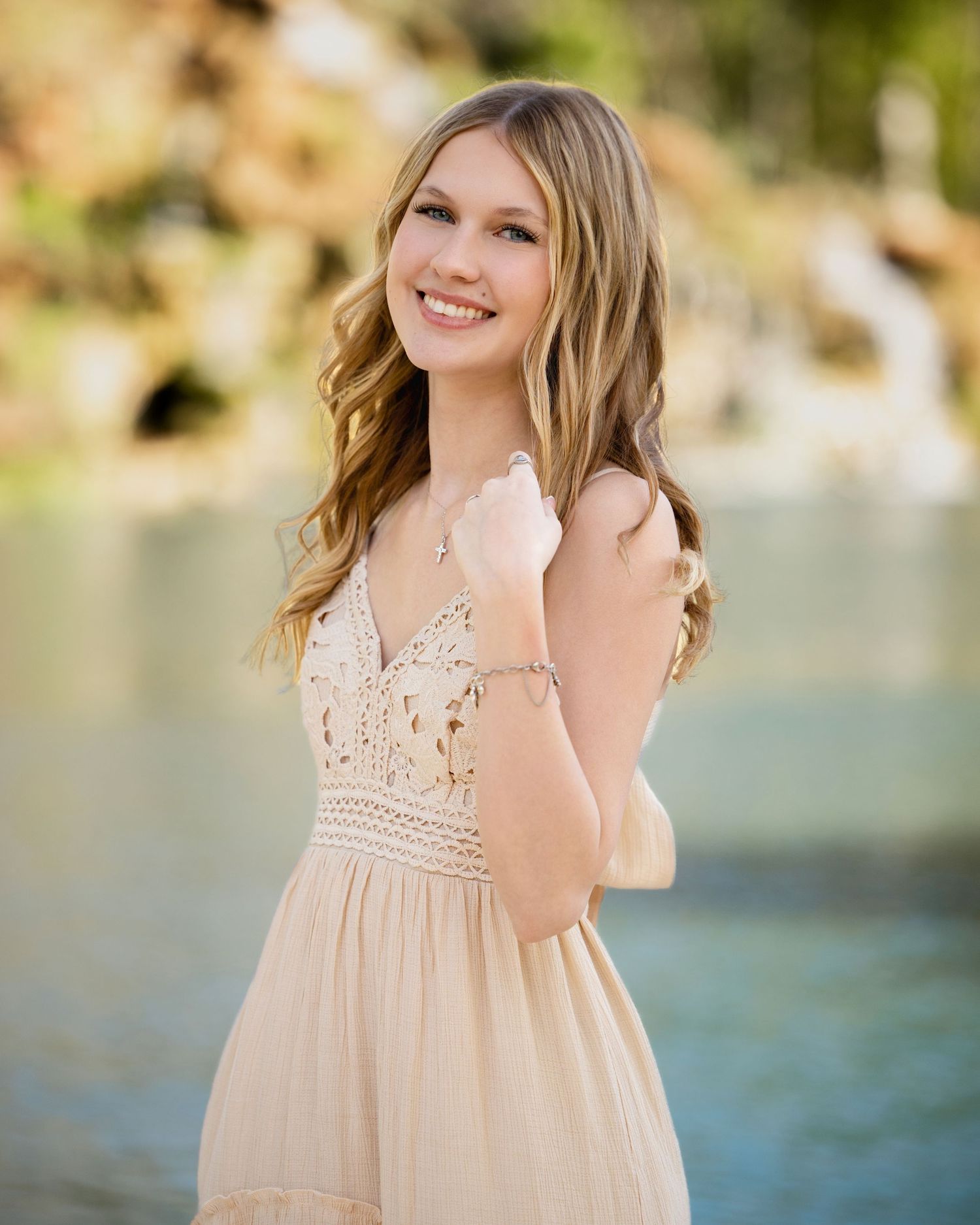 Young woman in a lace cream dress smiling outdoors near water, with wavy blonde hair and delicate jewelry.