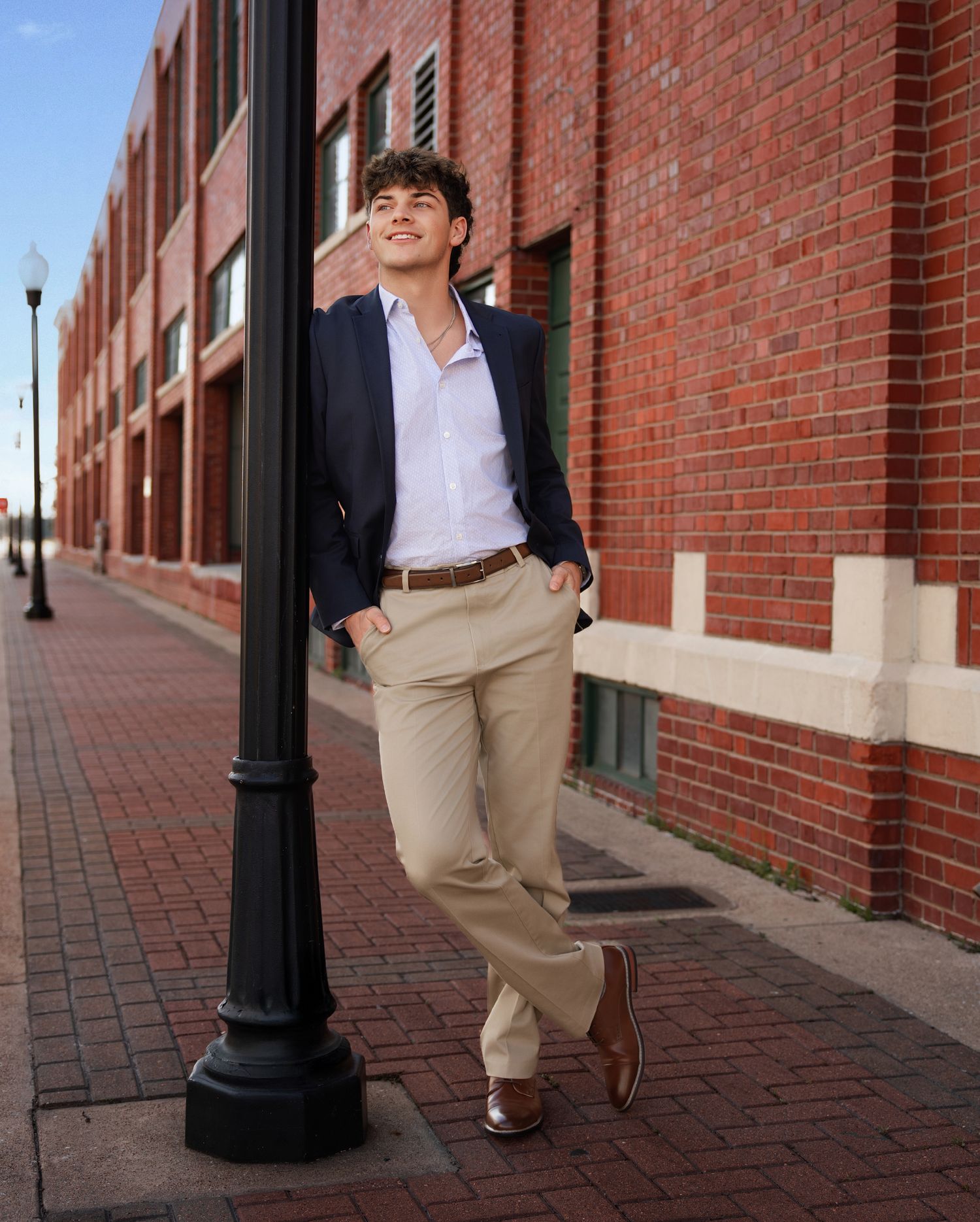 Young man in navy blazer and khakis leaning against a black lamppost on a brick sidewalk outside a red brick building.