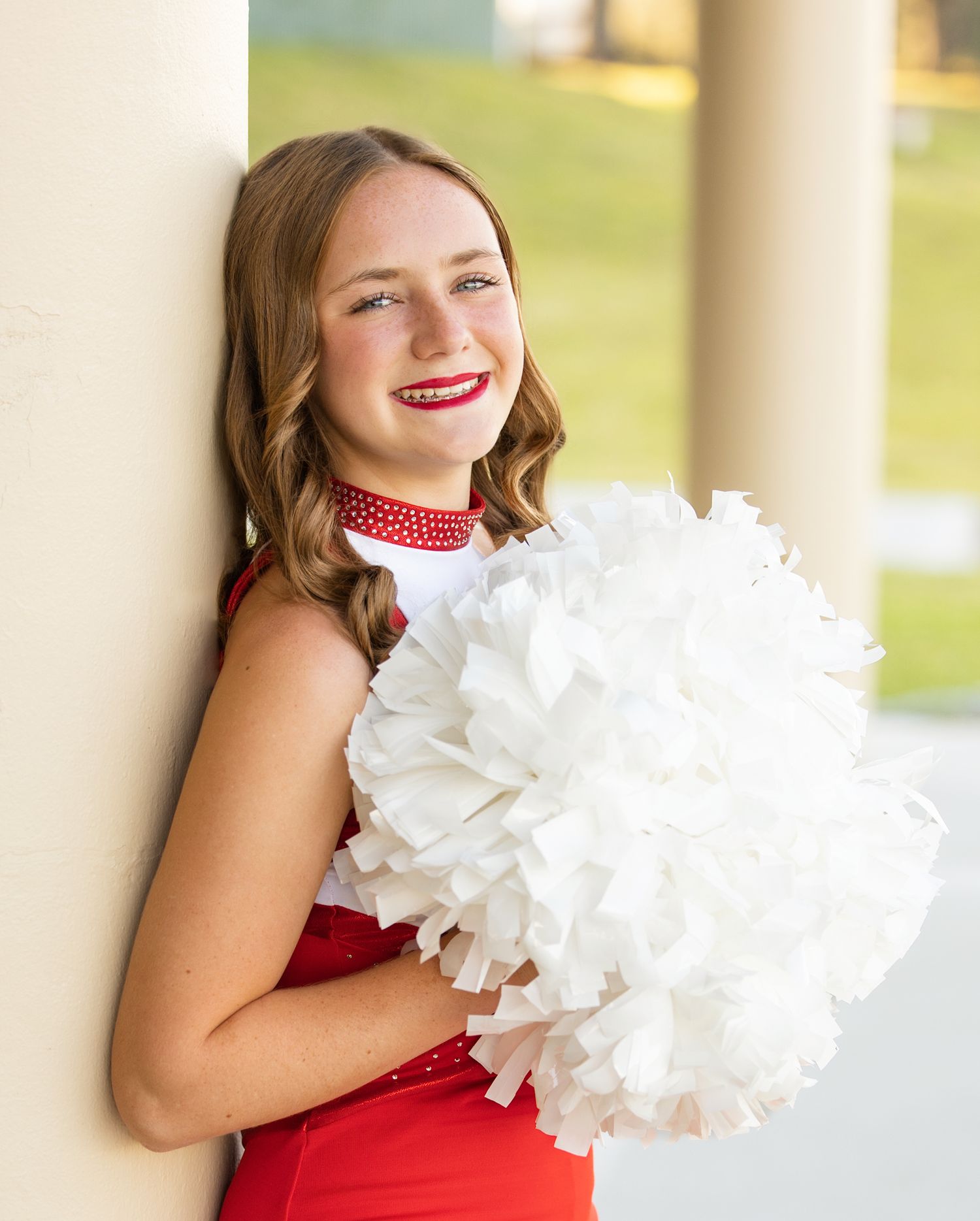 Cheerleader in red uniform holds white pom poms while posing against cream colored pillars outdoors.