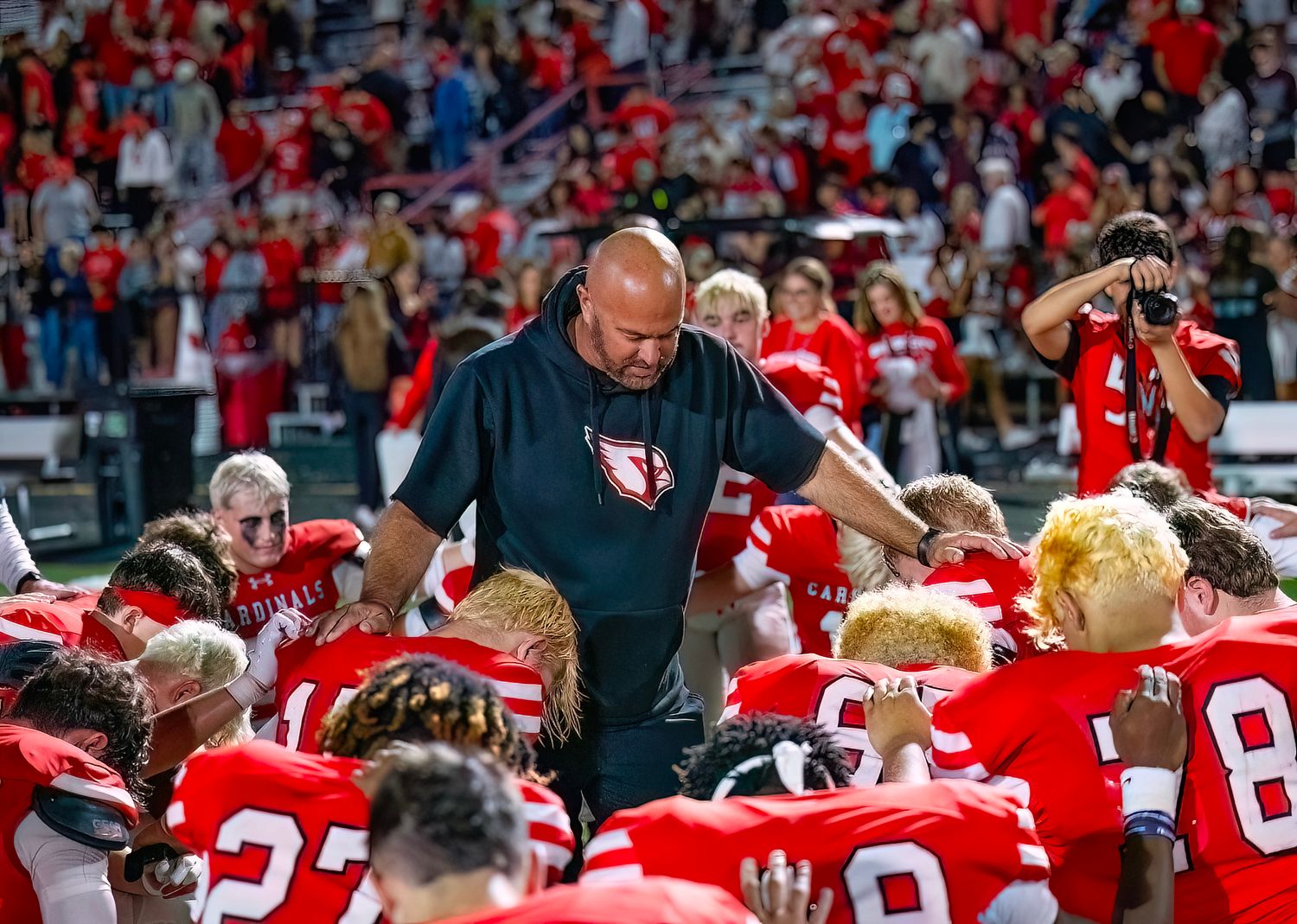 Athletic Director and team  in red jerseys celebrate and pray together in stadium seats during a live event.