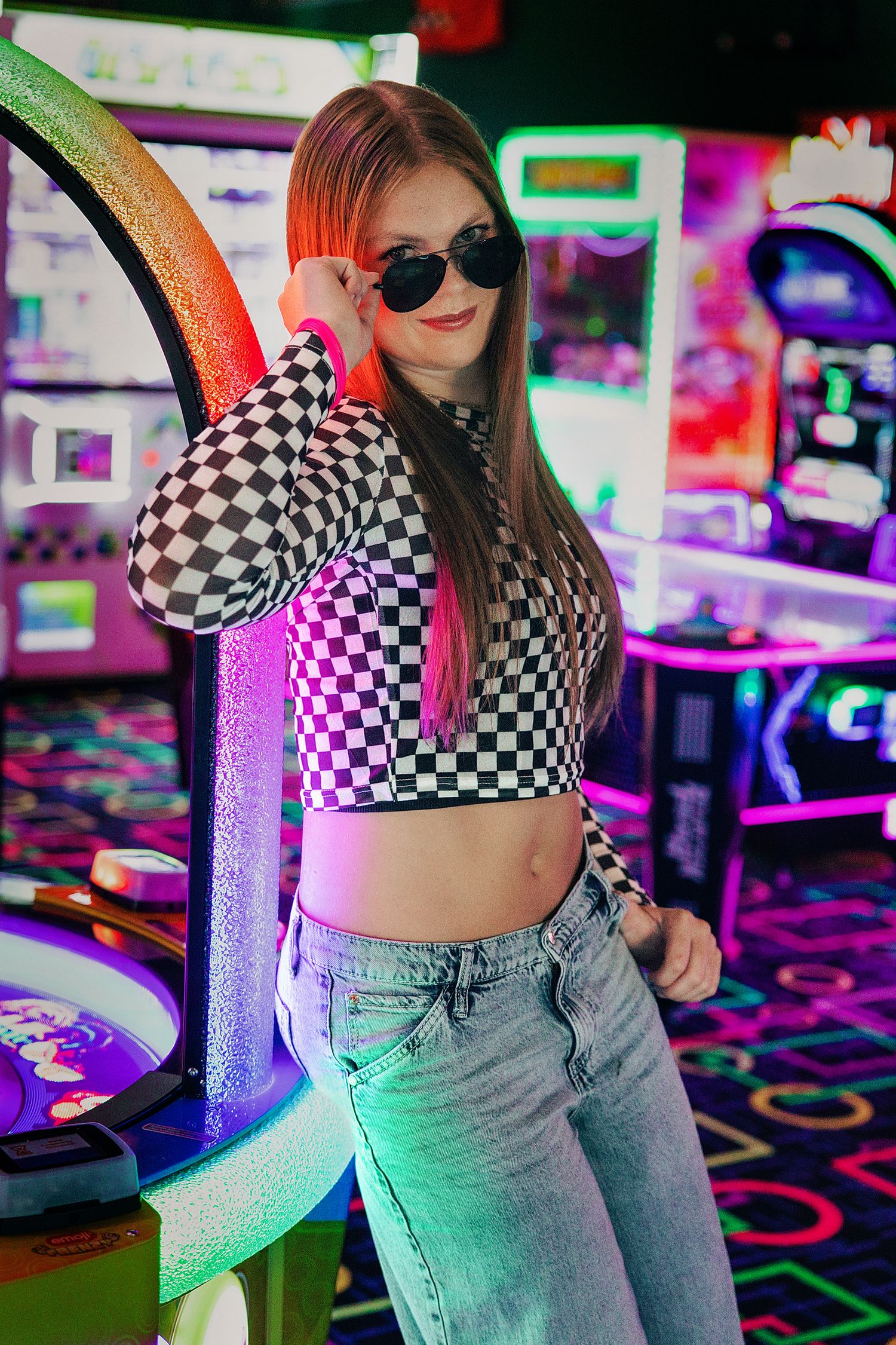 Young woman in checkered crop top and jeans poses at an arcade, wearing sunglasses under colorful neon lights.