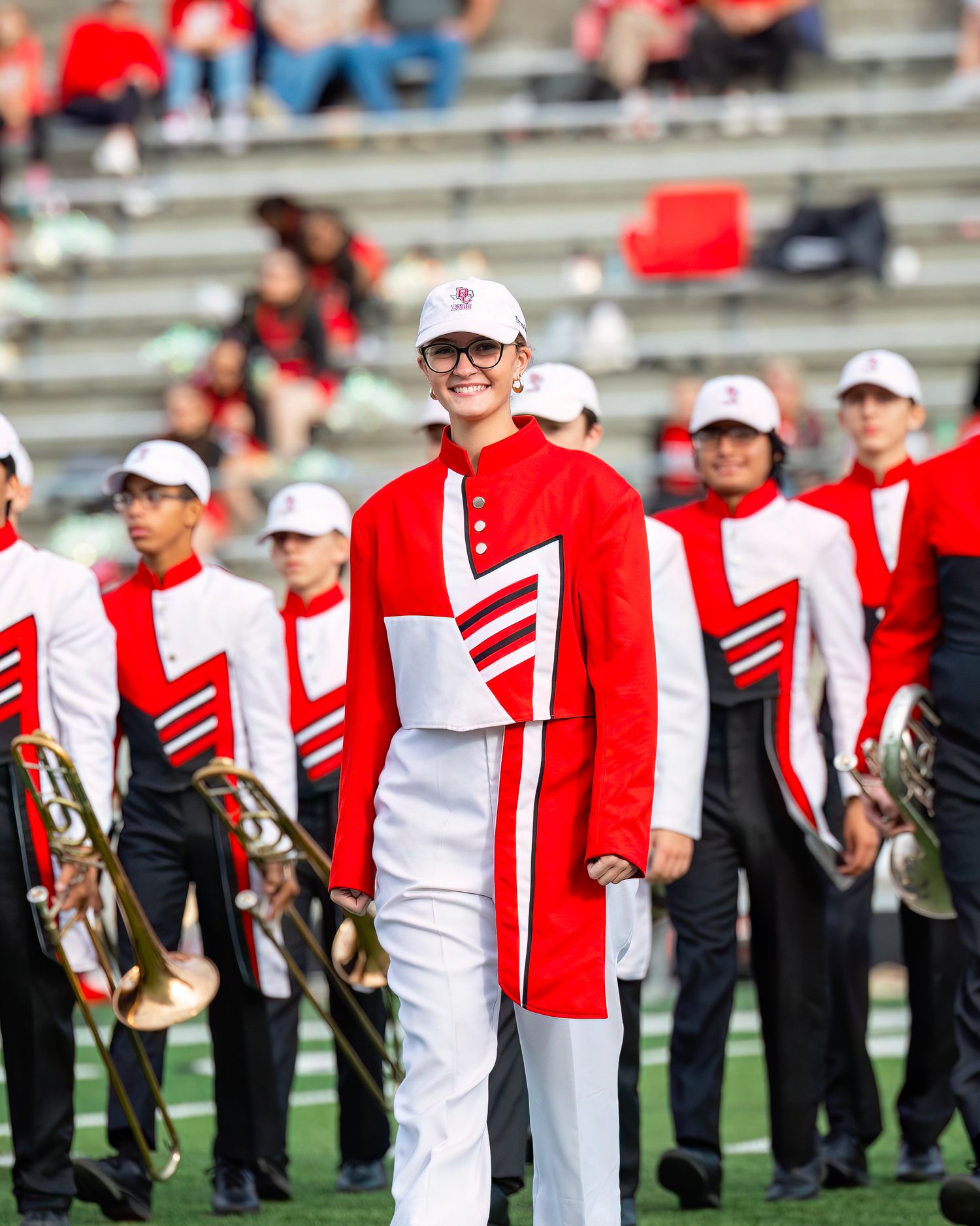 Marching band members in red and white uniforms stand in formation on a football field.