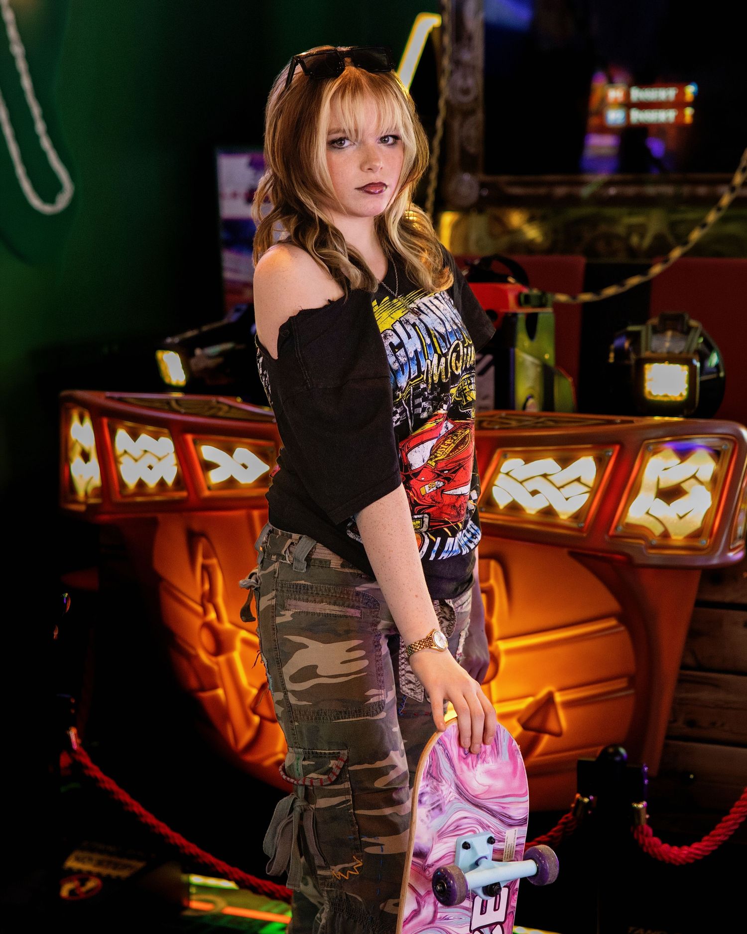 Young woman in camo pants and black off-shoulder top holding a pink skateboard in an arcade setting.