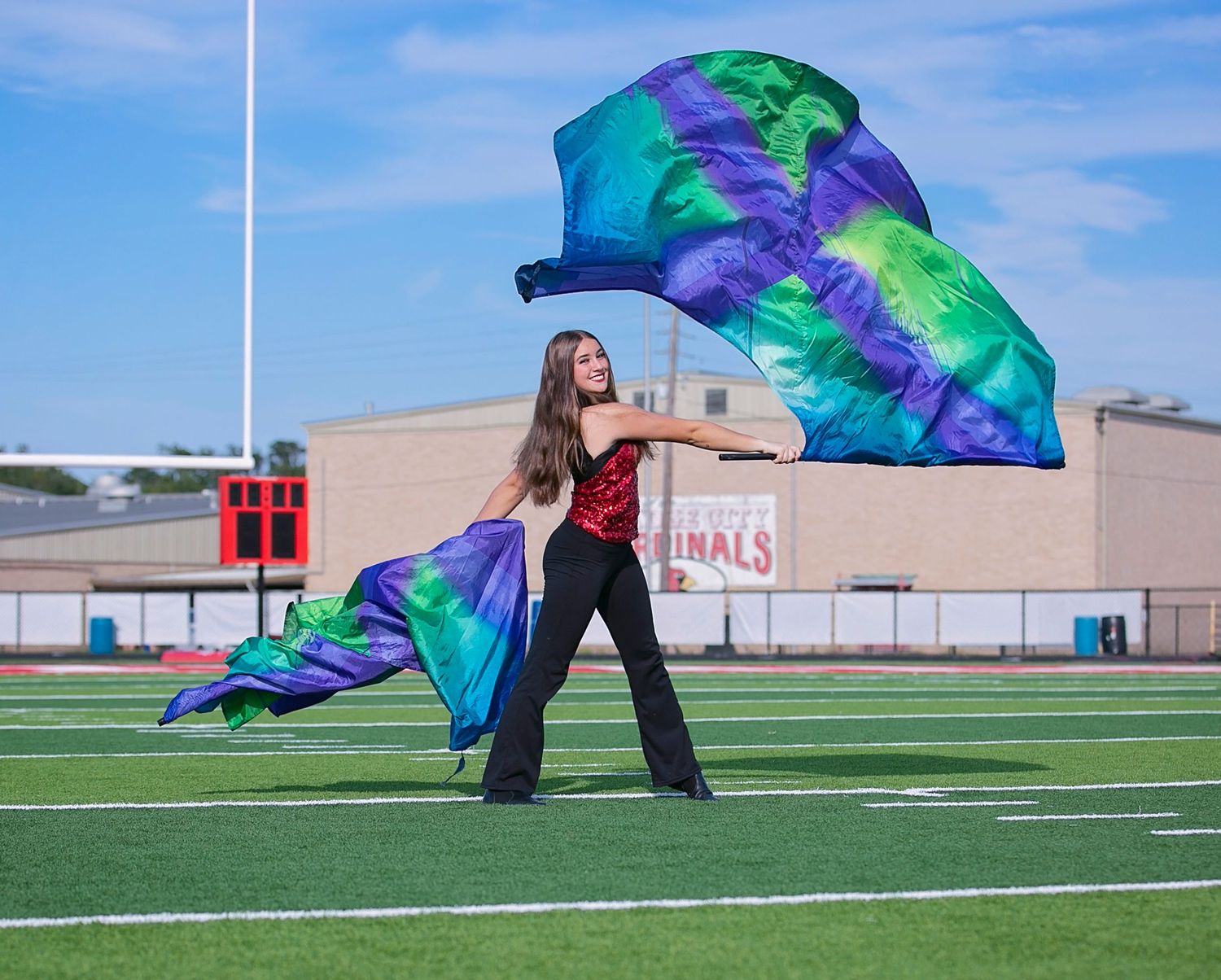 A person performs a flowing dance routine with a blue and green silk flag on a green athletic field under blue skies.