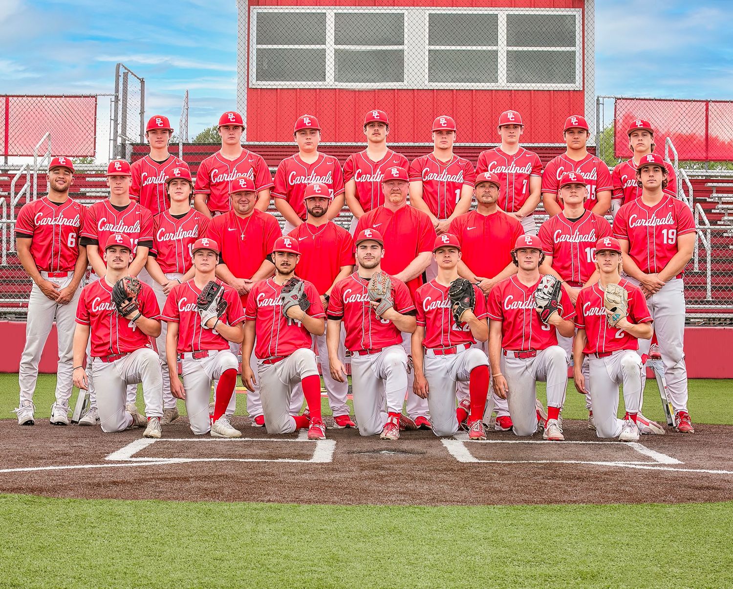 Baseball team wearing red jerseys kneels in formation on a field for their team photo.