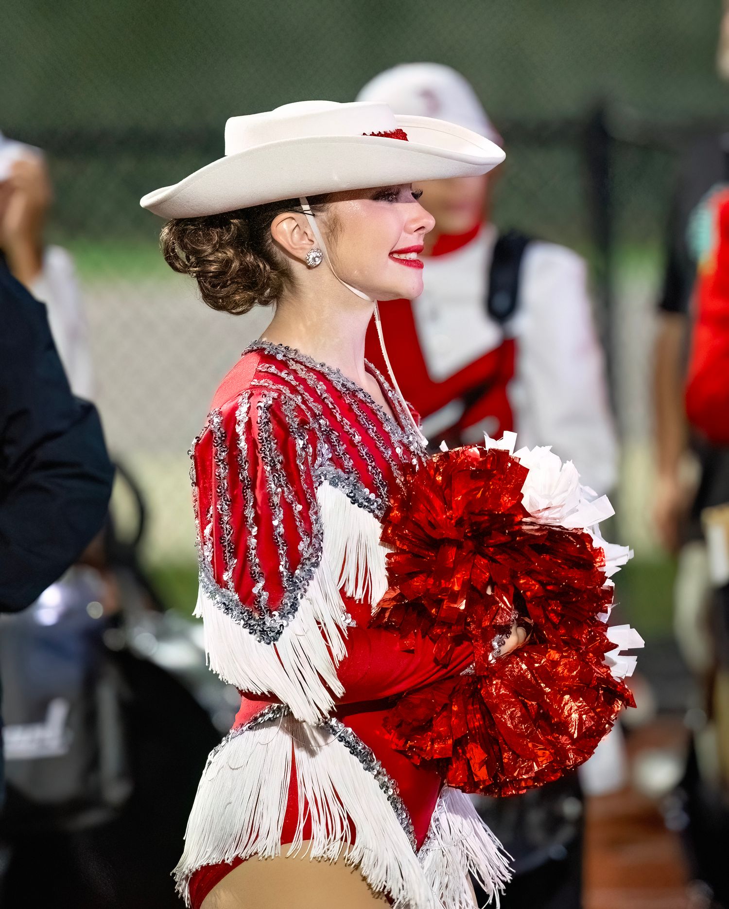 Performer in white hat and red sequined costume holds red pom poms during halftime show.