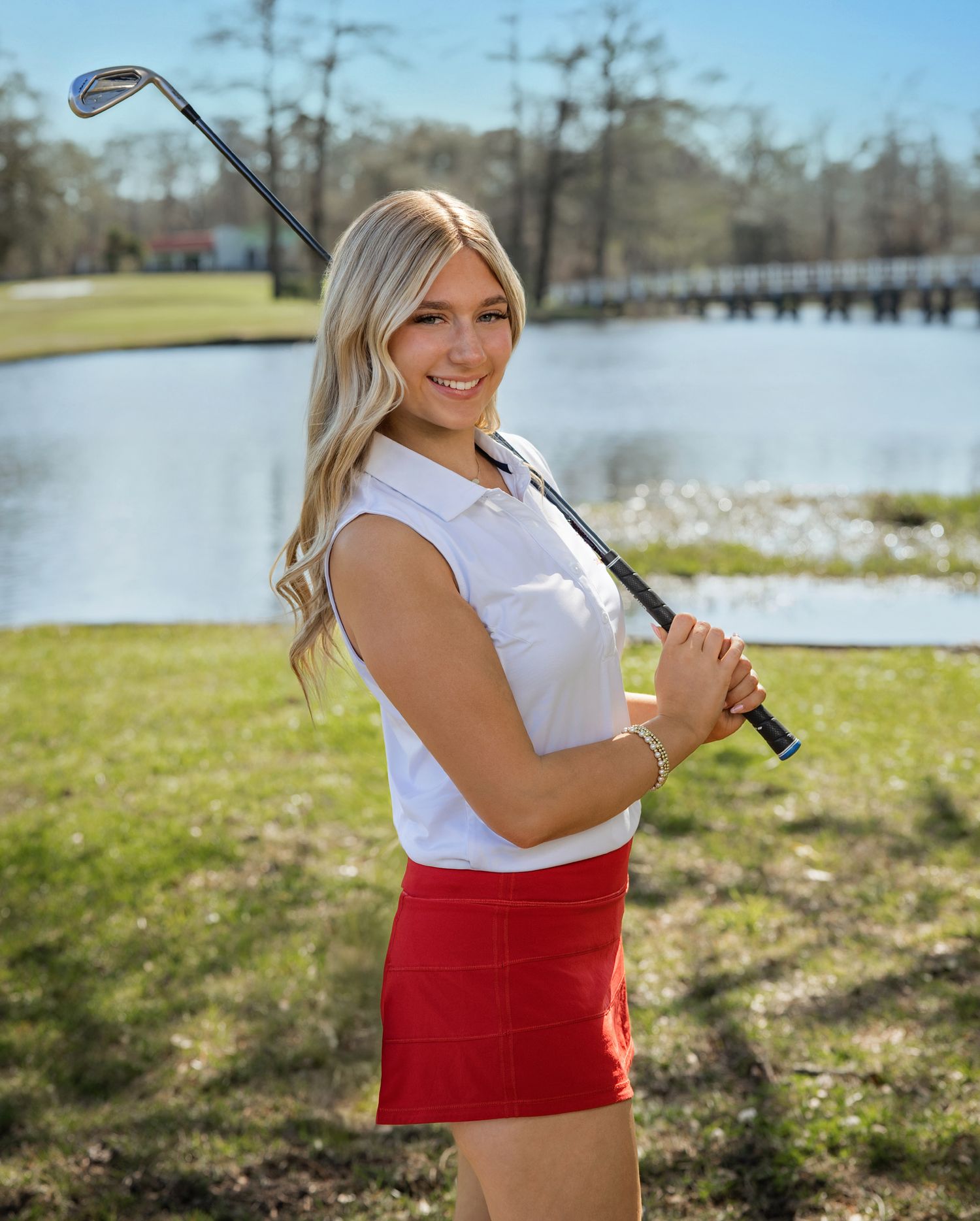 A person in a white top and red skirt poses with a golf club on a sunny day near a lake.