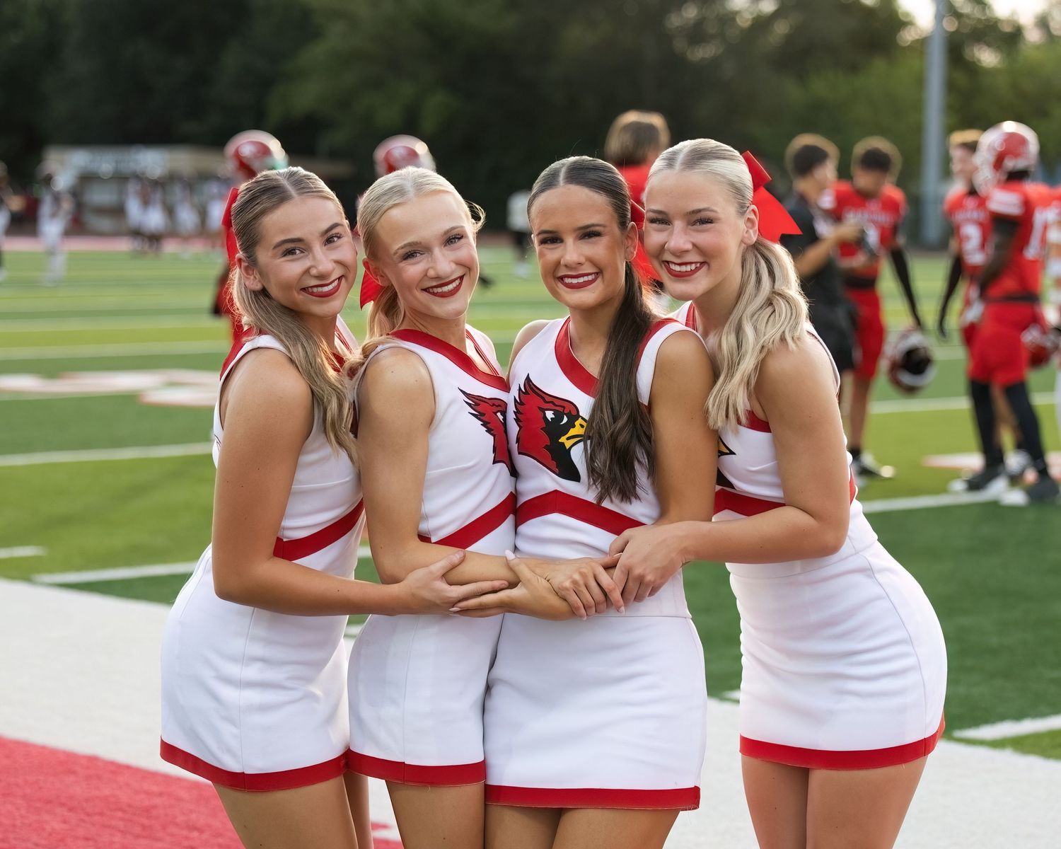 Cheerleaders in red and white uniforms embrace and smile while posing together on a sports field during practice.
