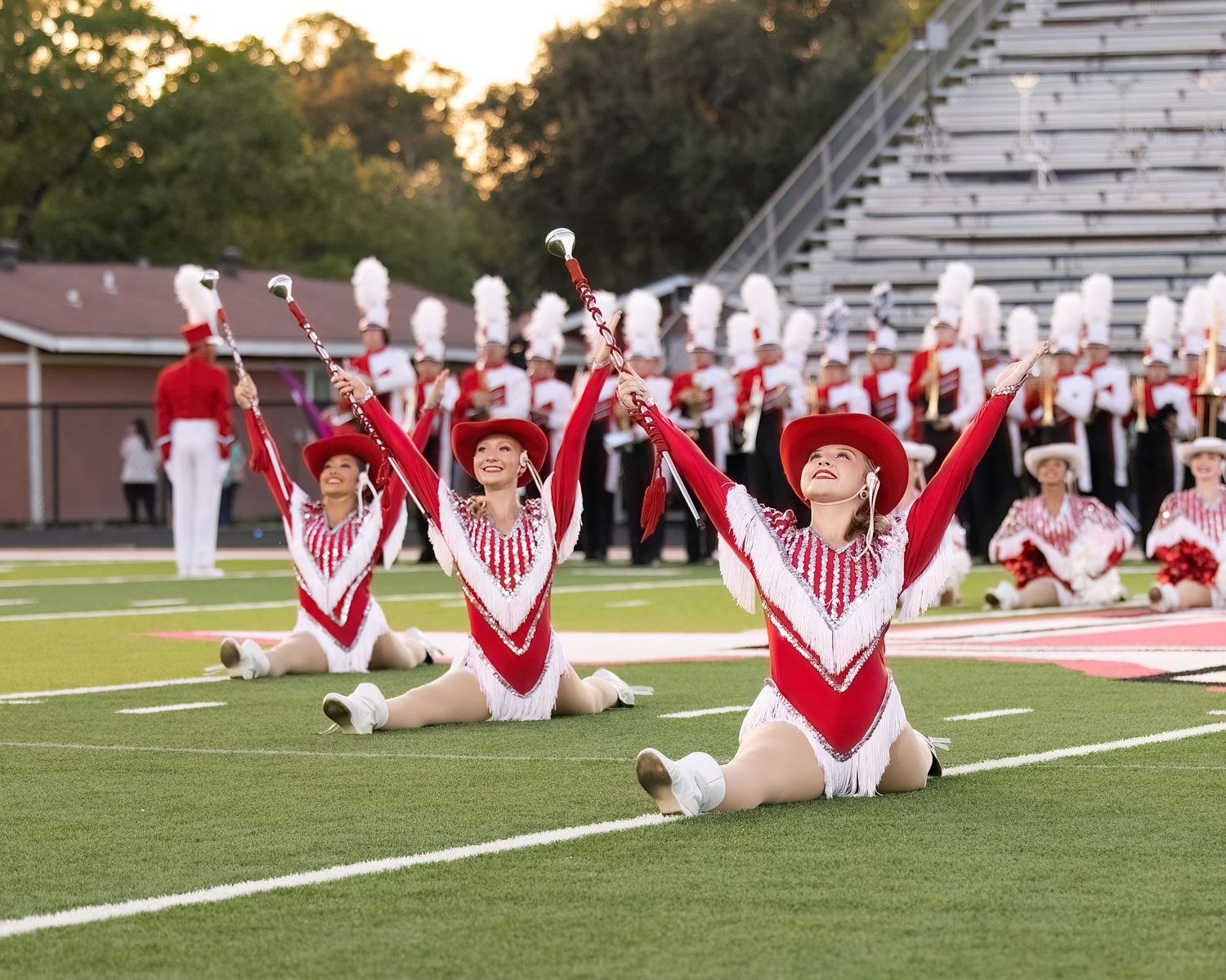 Cheerleaders in red and white uniforms perform choreographed formations on a football field at sunset.