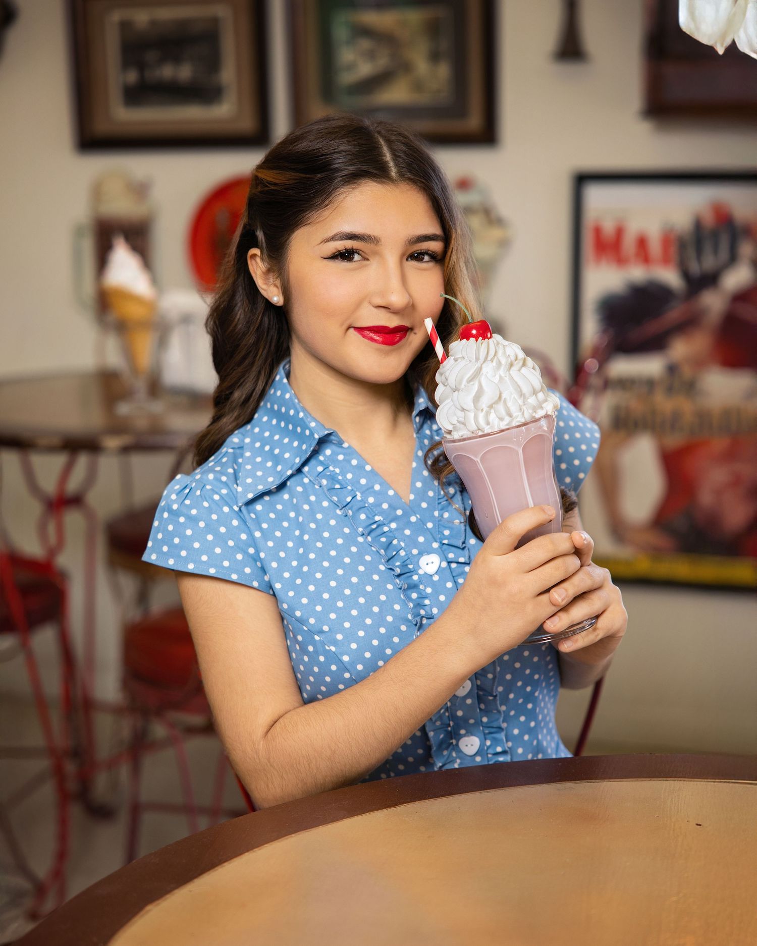 Person in blue polka dot dress sitting at diner counter holding pink milkshake.