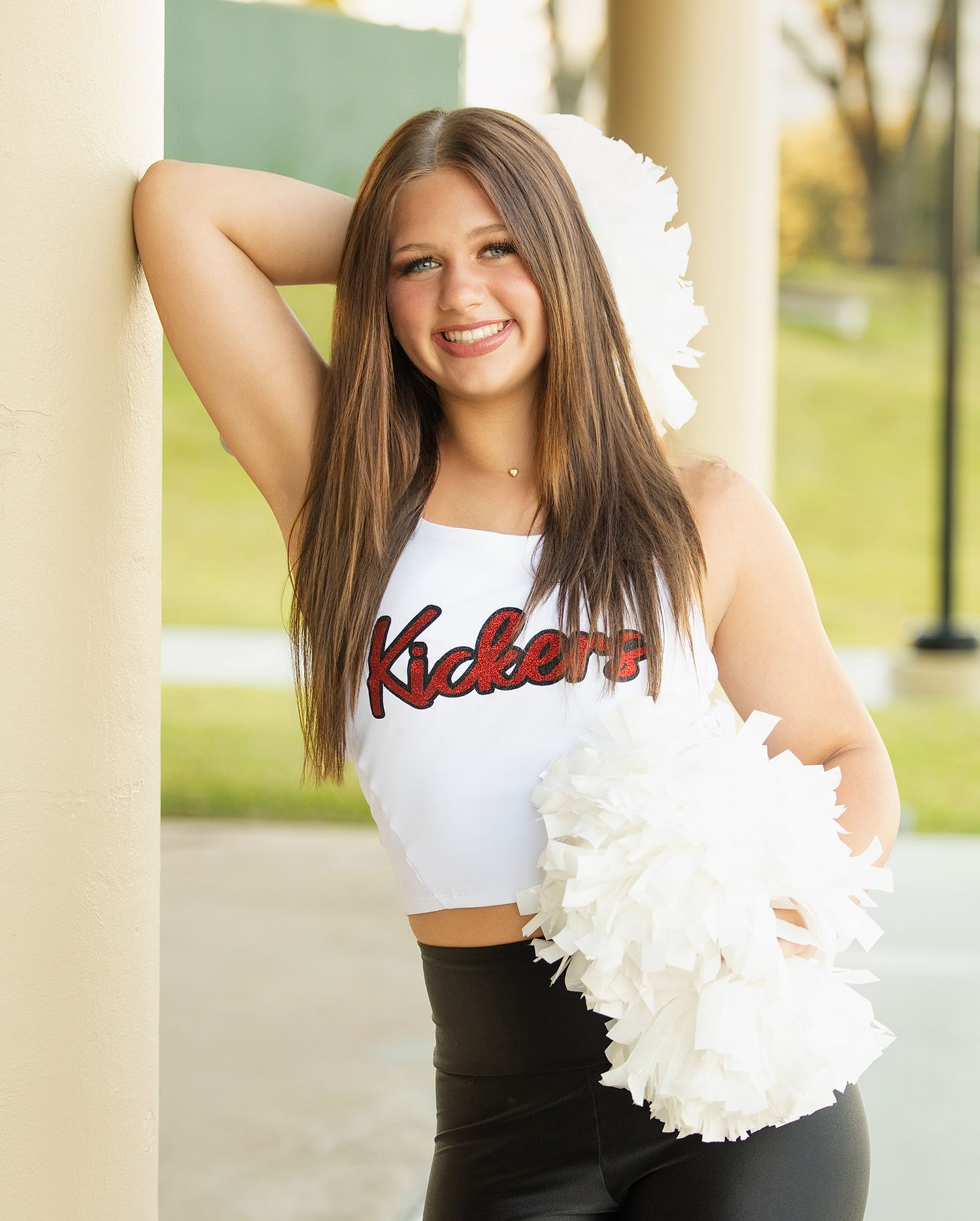 Cheerleader in white and black uniform holds white pom poms while posing between columns.