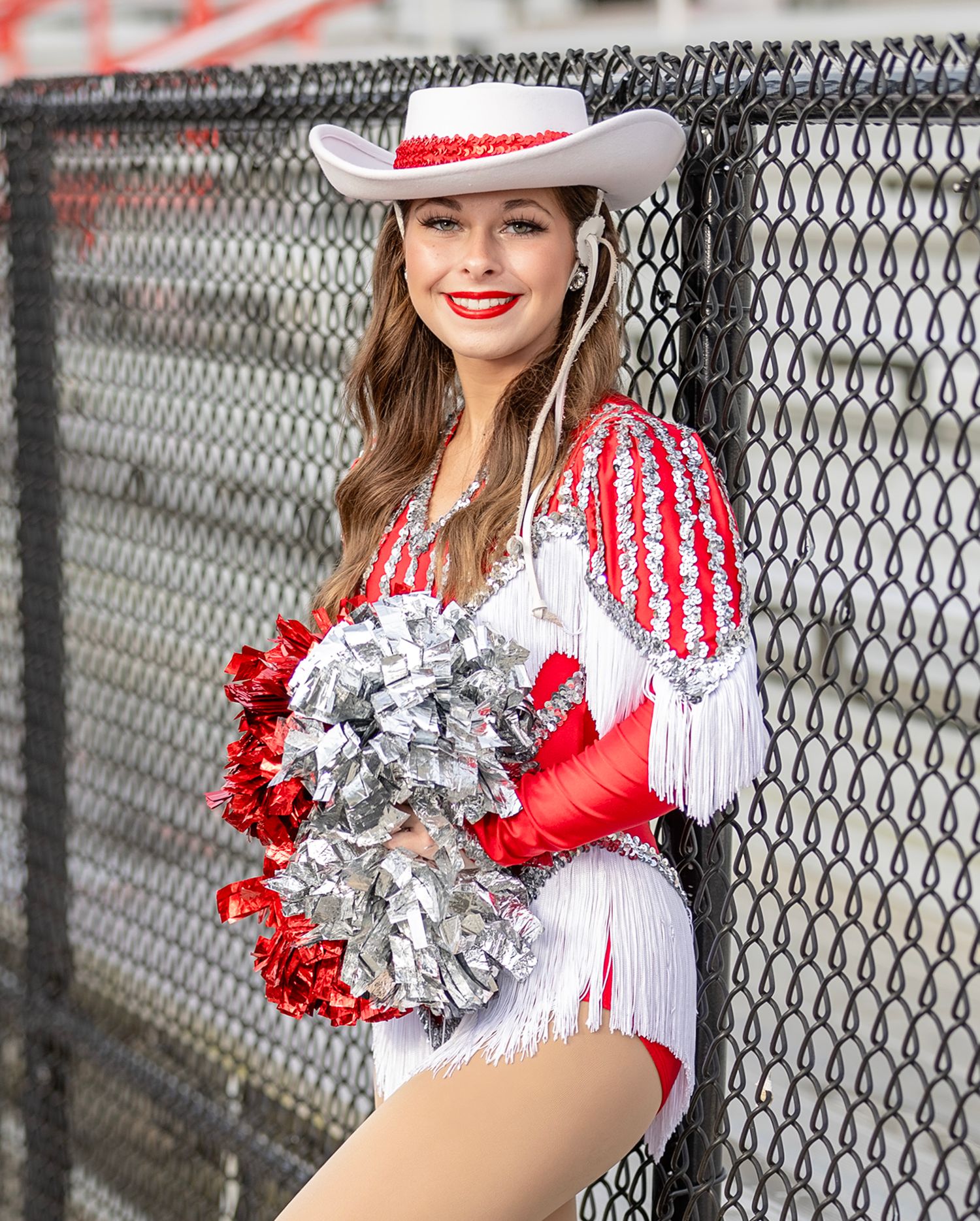Cheerleader in red and white uniform with silver pom poms poses against chain link fence.