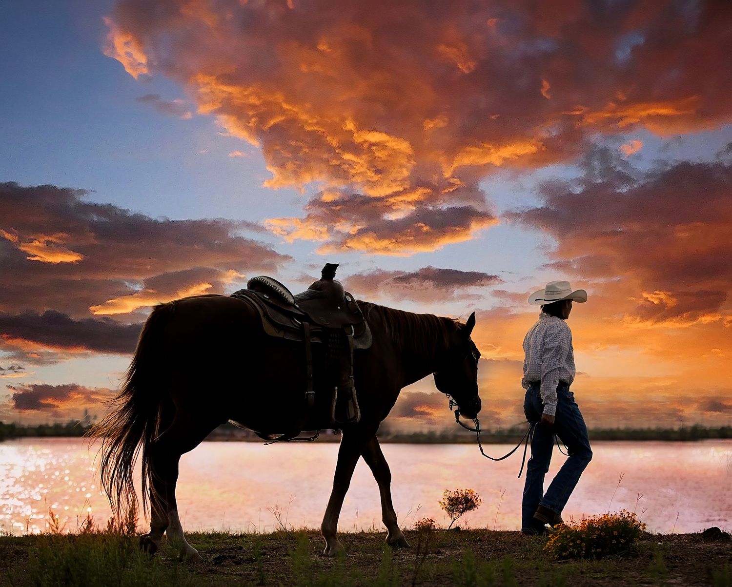 Cowboy leading a dark horse by reins at sunset with dramatic orange and red cloudy sky reflecting on water.