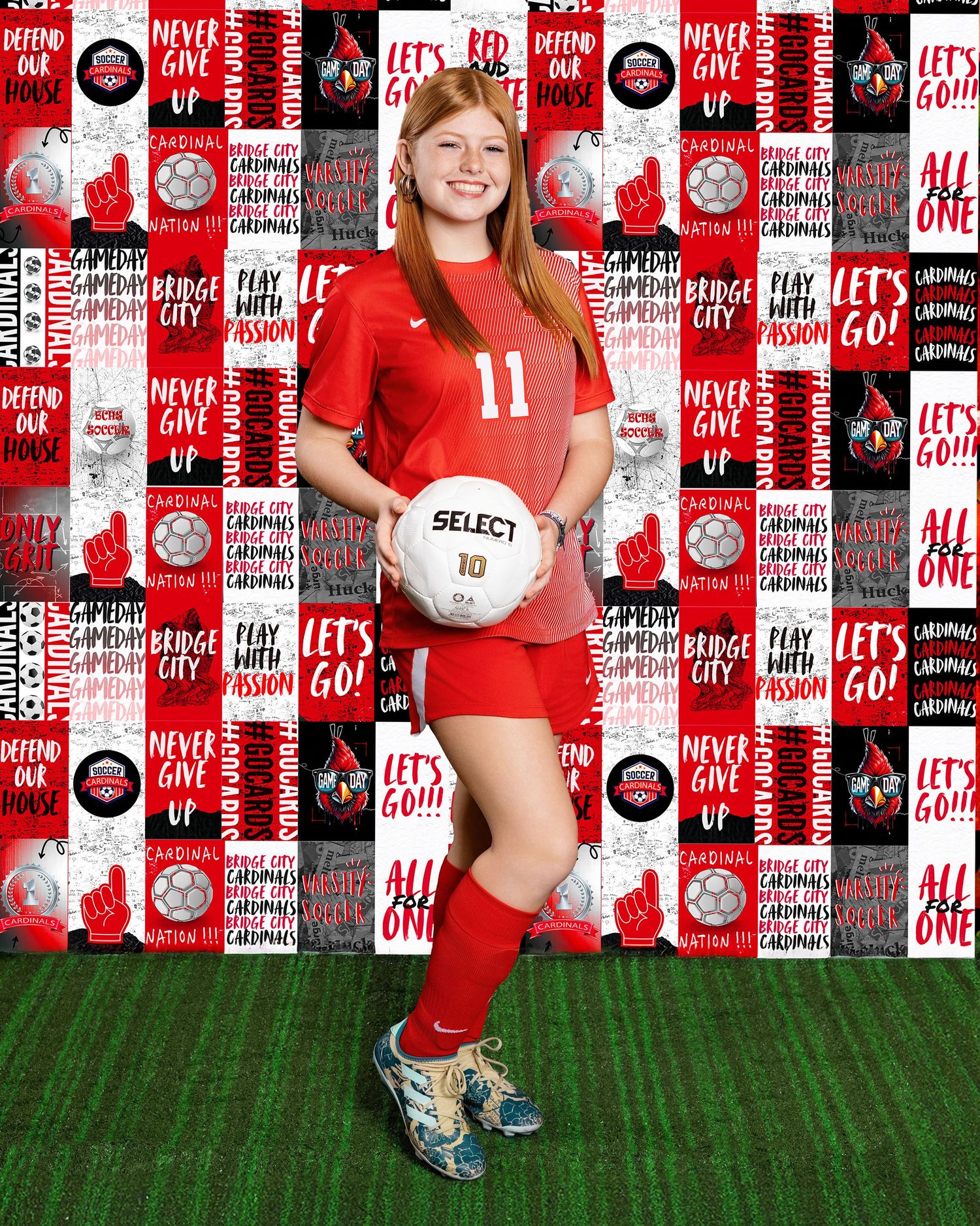 Soccer player in red uniform stands on turf field with ball against branded backdrop.