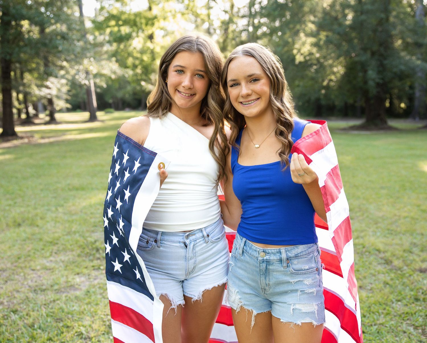 Two people draped in American flags pose together outdoors in summer clothing at a park.