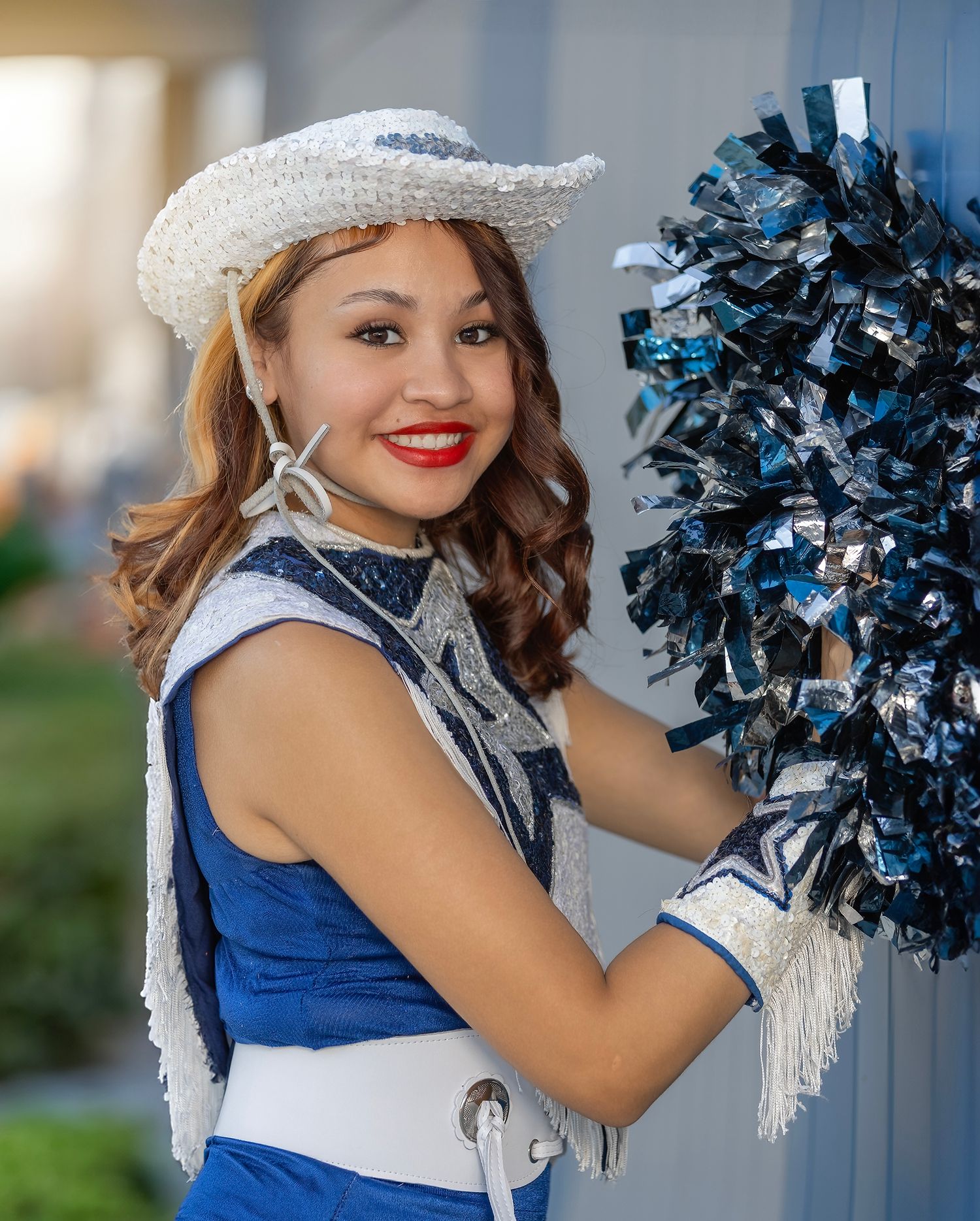 Cheerleader in blue uniform and white hat holds silver pom poms while posing for photos outdoors.