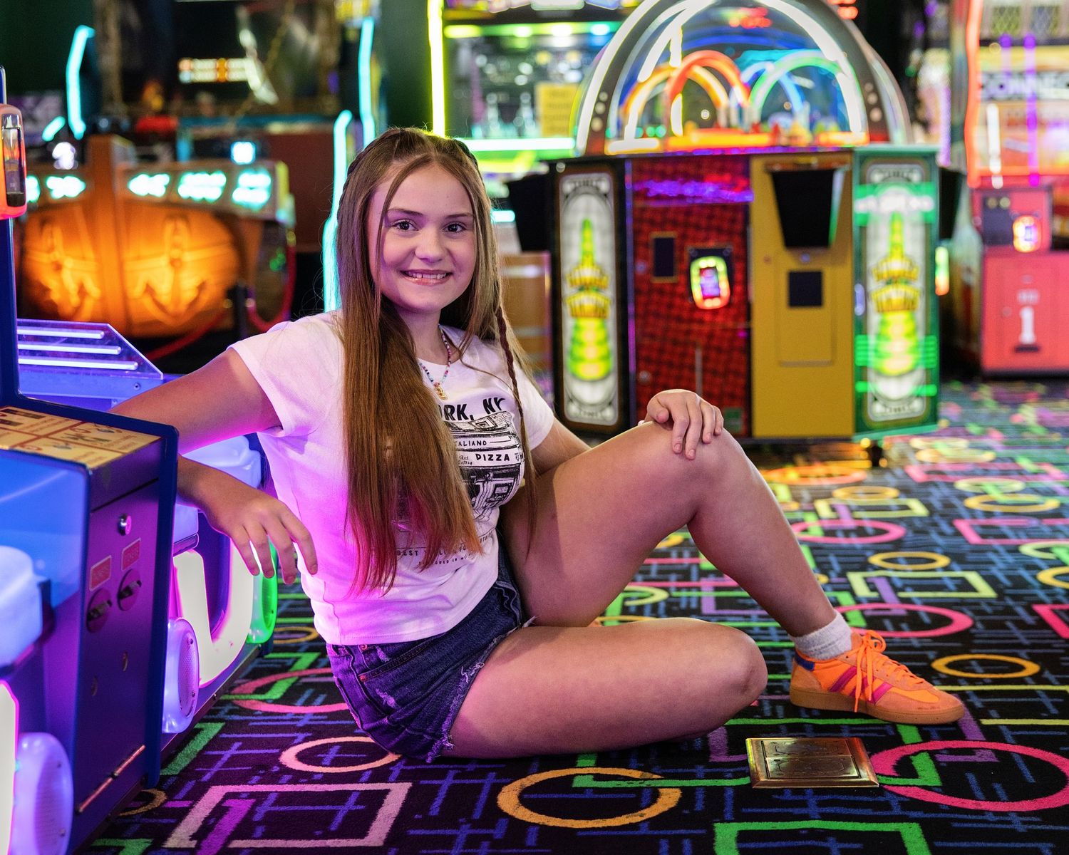 Young woman sitting on colorful carpet in an arcade, smiling, with neon lights and game machines behind her.