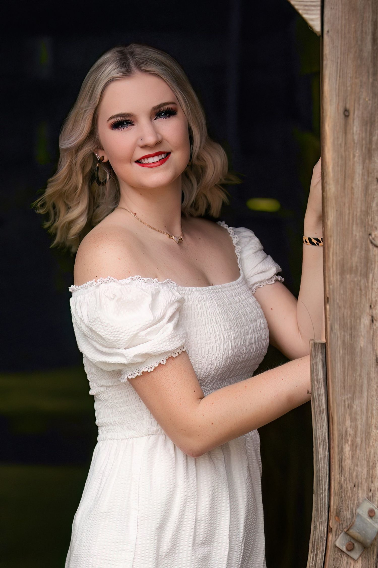Young blonde woman in white off-shoulder dress smiling beside rustic wooden door outdoors.