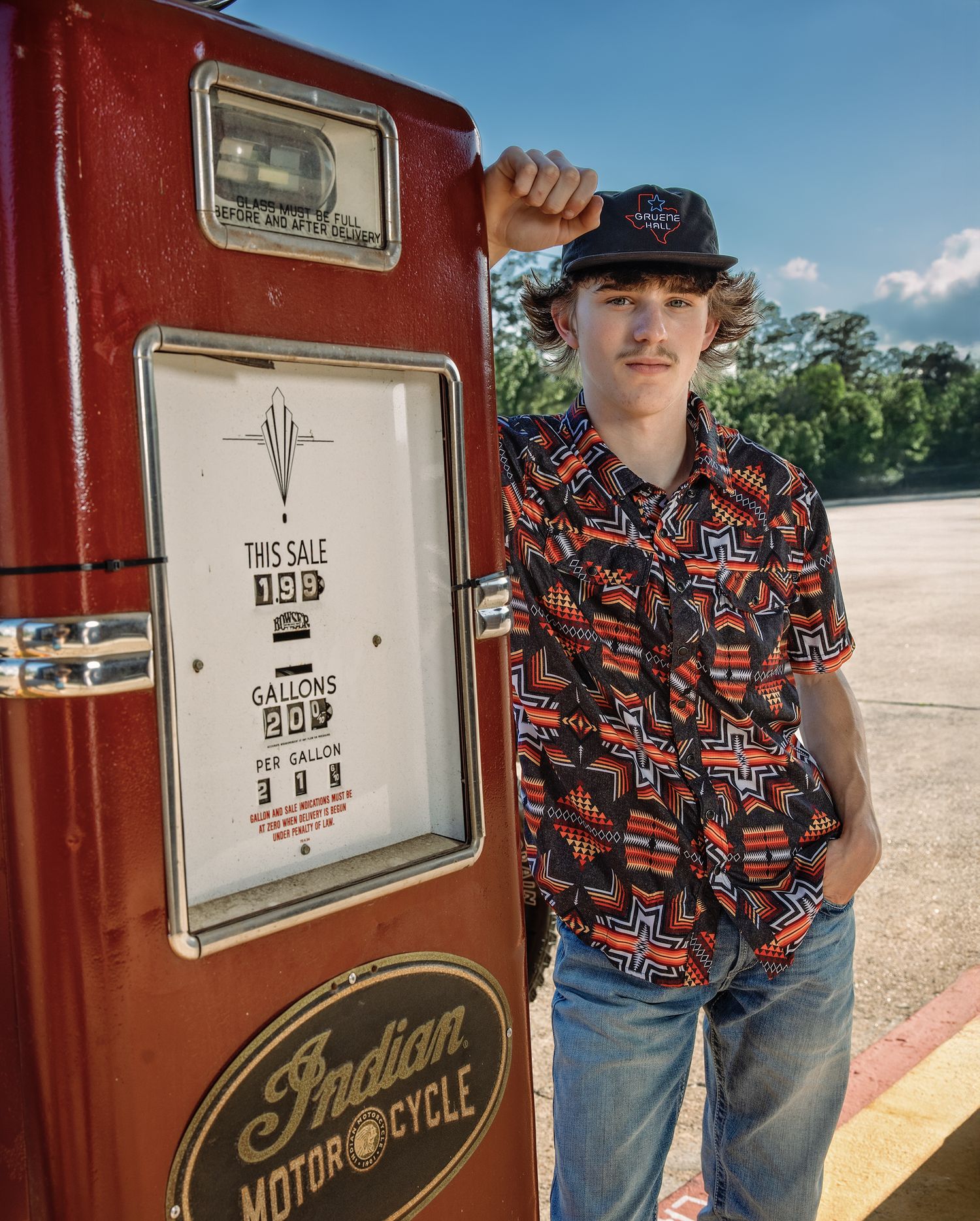 Soon to be graduate in a patterned shirt stands next to a vintage red Indian Motorcycle Oil vending machine.