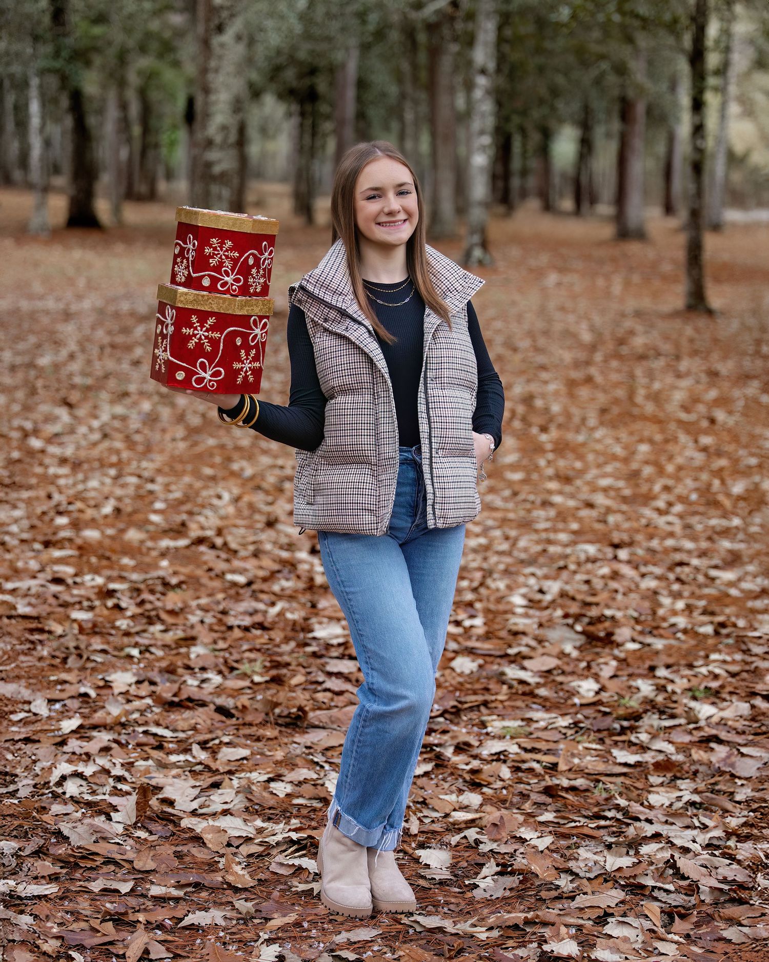 Person in plaid vest and jeans holds red gift boxes while standing in autumn forest covered with fallen leaves.