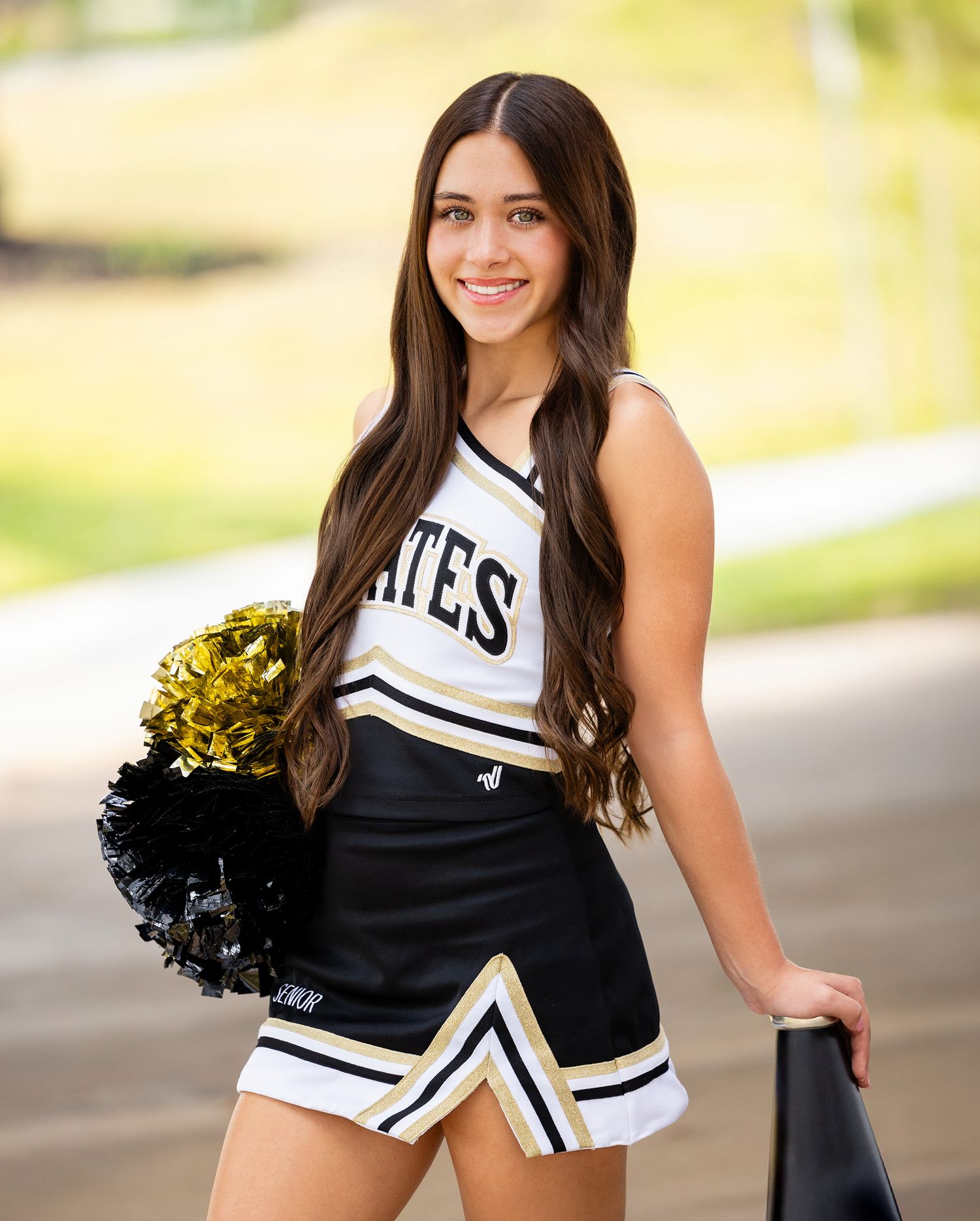 VHS cheerleader in black and white uniform performs a dynamic pose with pom poms against a sunny outdoor background.