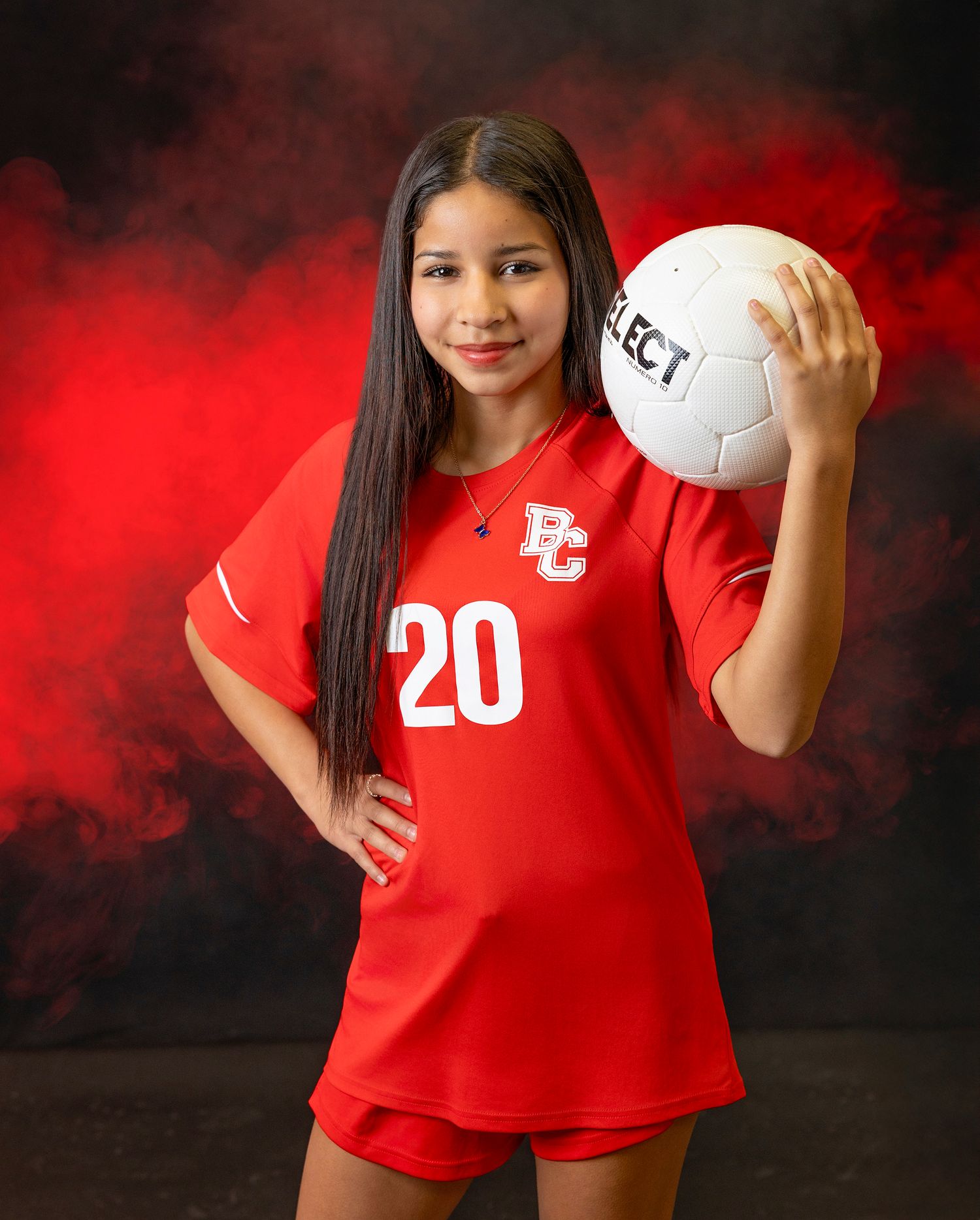 Soccer athlete in red uniform number 20 holds white ball against red smoky backdrop.