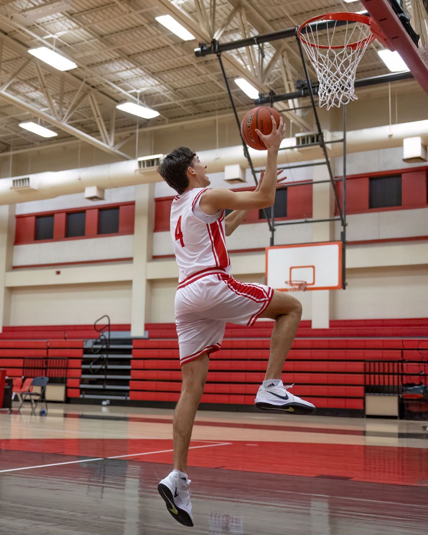 Basketball player in white and red uniform performs a layup shot in a high school gymnasium with red bleachers.